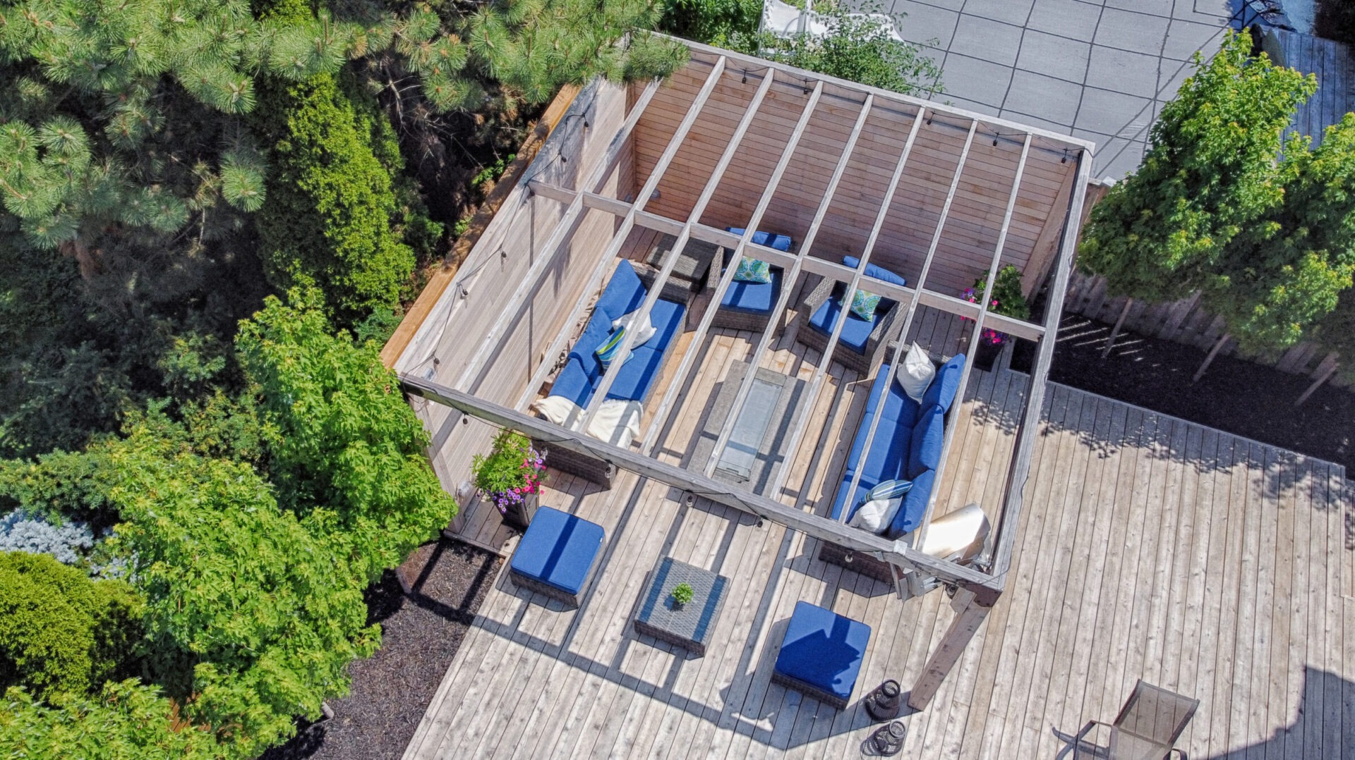 An aerial view of a wooden deck patio with modern outdoor furniture, surrounded by lush greenery and a concrete walkway in the distance.