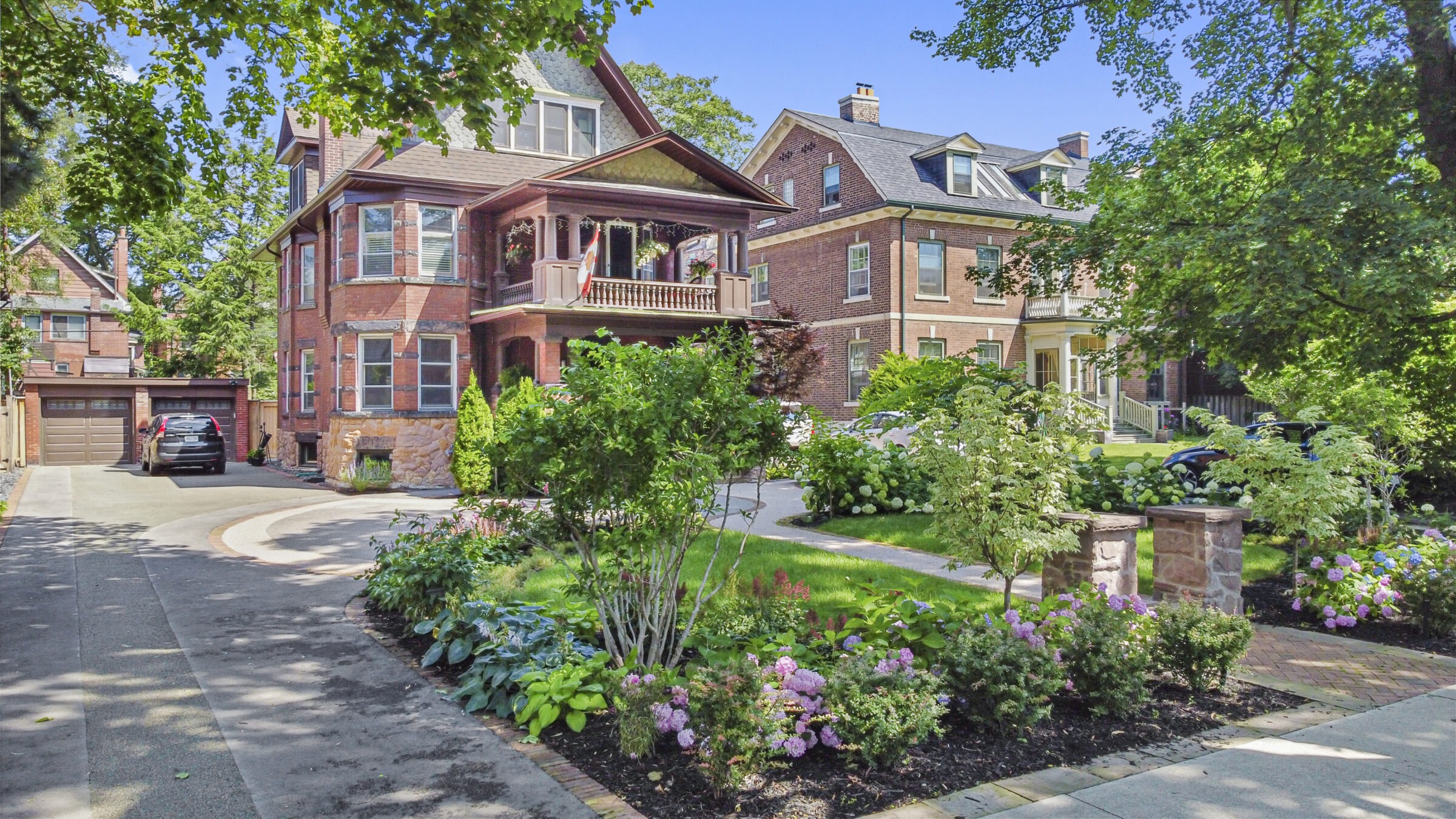 A sunny residential street with large, elegant brick houses, lush gardens, a curved driveway, and mature trees under a clear blue sky.