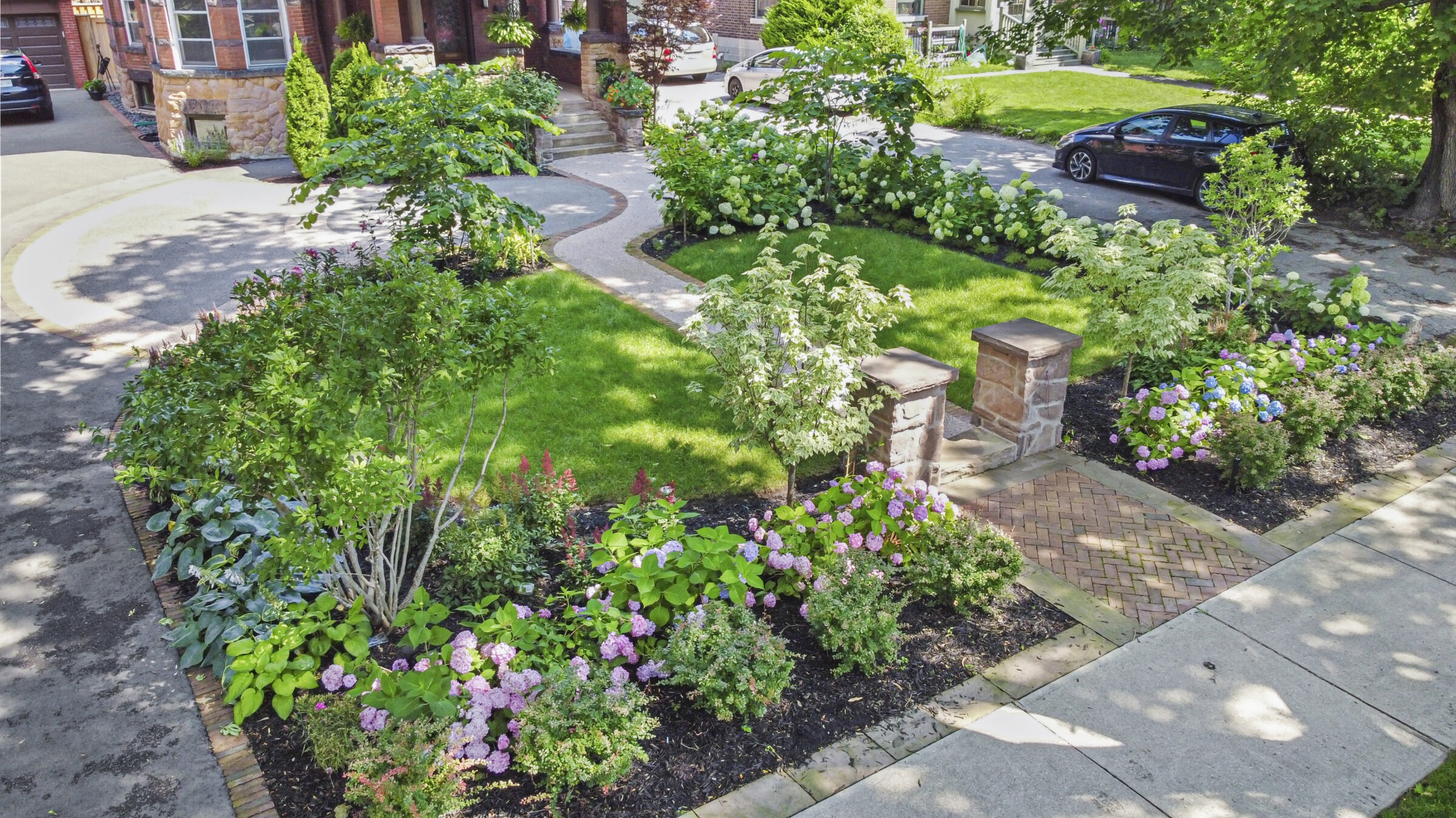 Lush front yard garden with flowering shrubs, green lawn, stone pathway, trees, and a parked car beside a house with a brick facade.