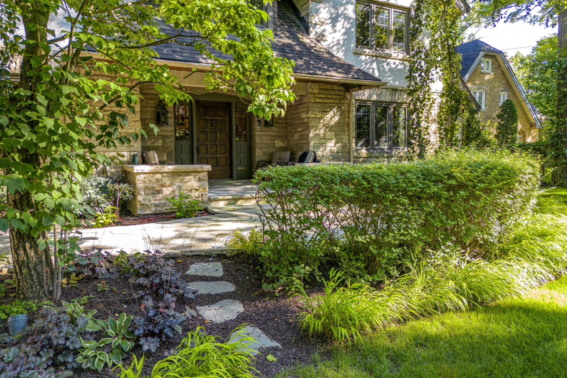 A stone house with lush green landscaping, stepping stones leading to the entrance, surrounded by trees and shrubs on a sunny day.