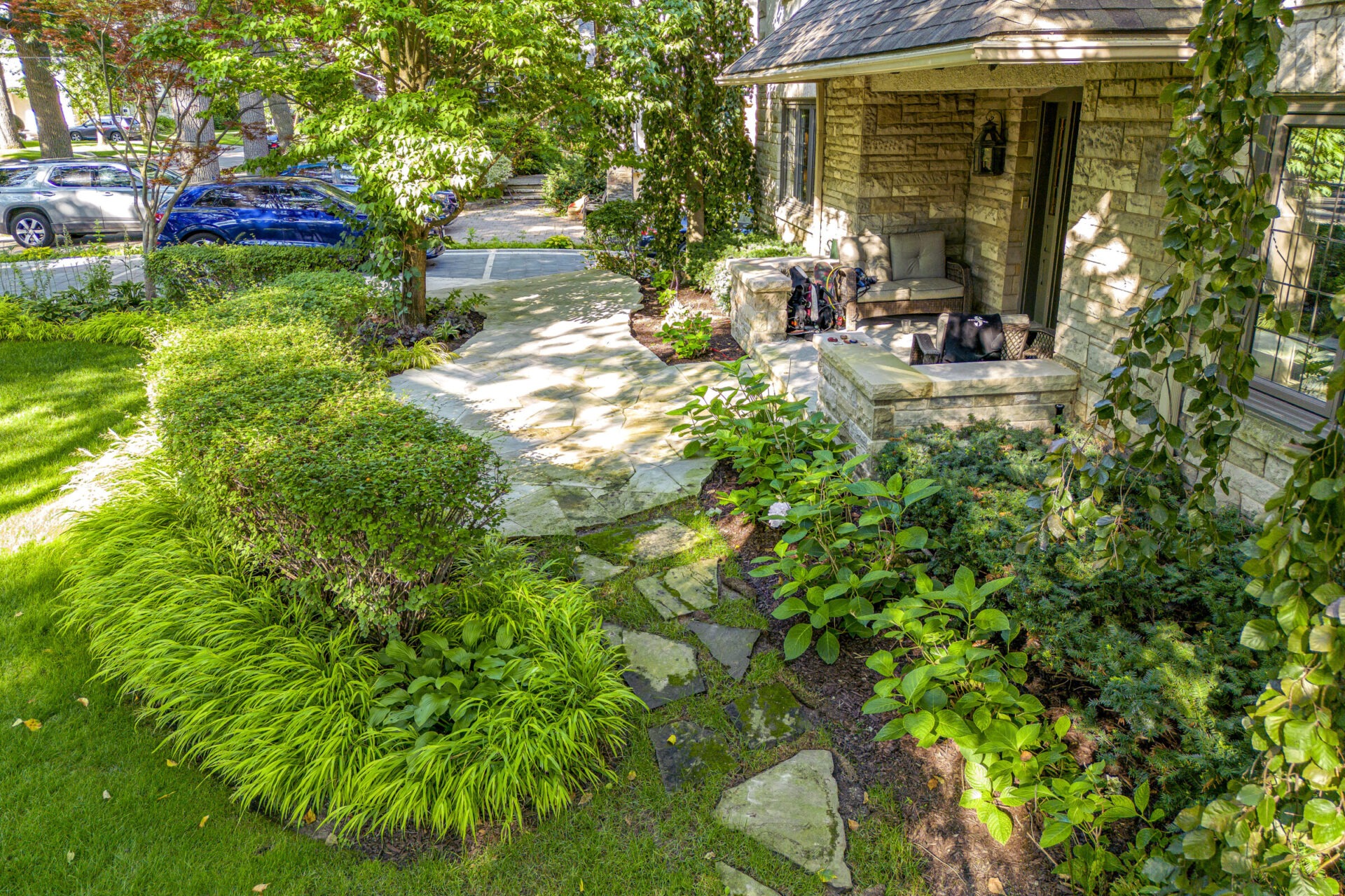 A lush garden with a stone pathway leading to a house with a sitting area where a person and a dog relax. Greenery surrounds the serene scene.