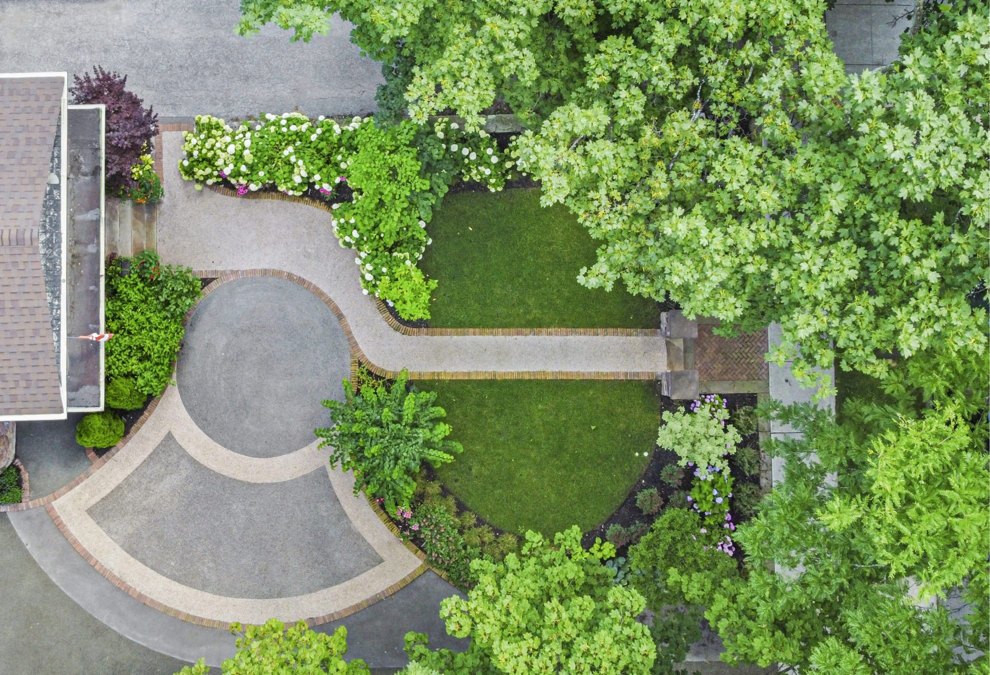Aerial view of a landscaped garden with winding pathways, lush greenery, trees, and flowering shrubs near a section of a house's roof.