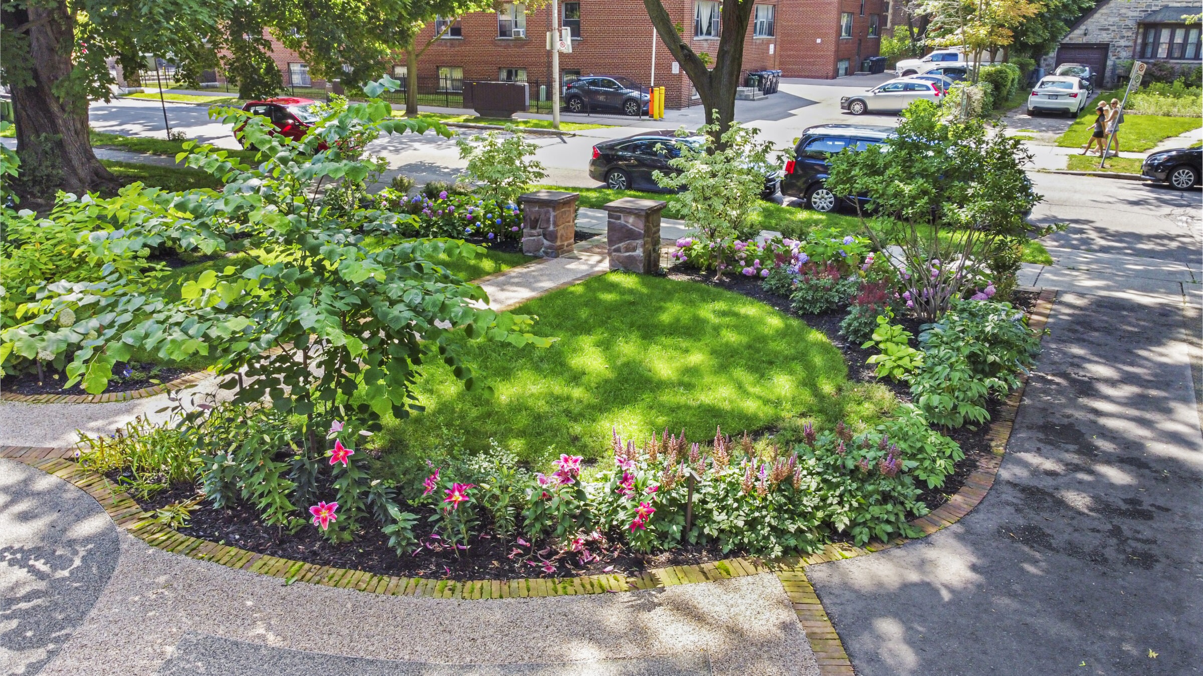 A vibrant garden with lush green grass, diverse plants, and a paved walkway, surrounded by urban environment, with buildings and passing pedestrians.
