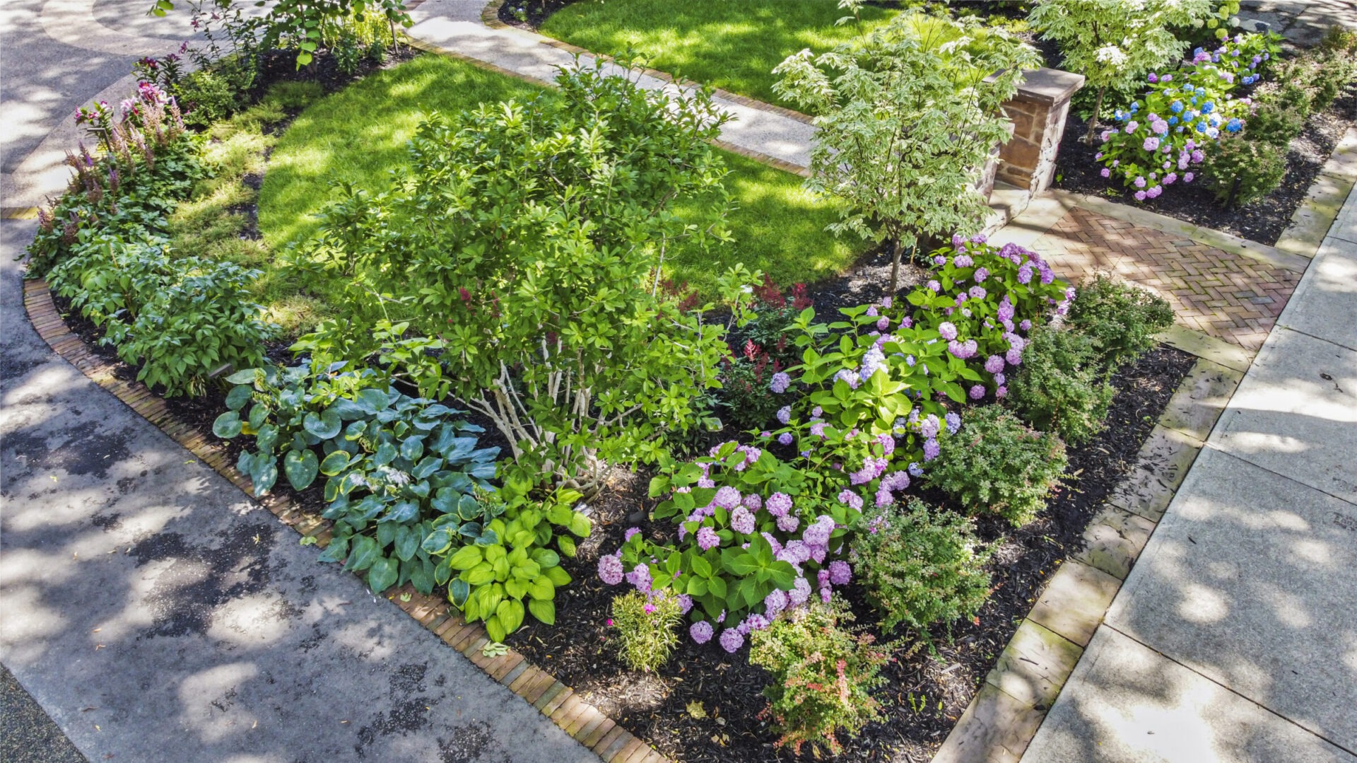 A well-maintained garden path with brick edging, surrounded by lush greenery, flowering shrubs, and trees under a clear sky, suggesting a serene landscape.