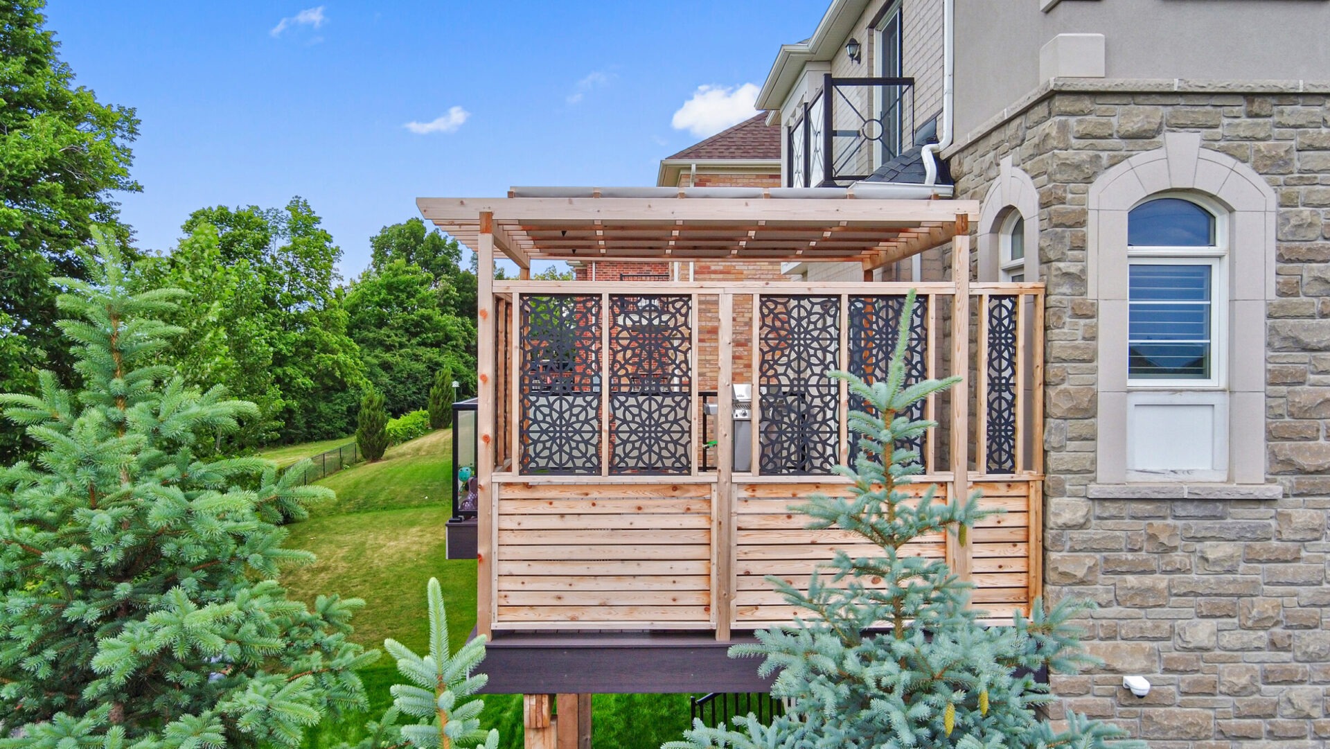 This image depicts a modern house with a stone facade and a wooden deck featuring intricate black privacy screens, set against a backdrop of greenery.