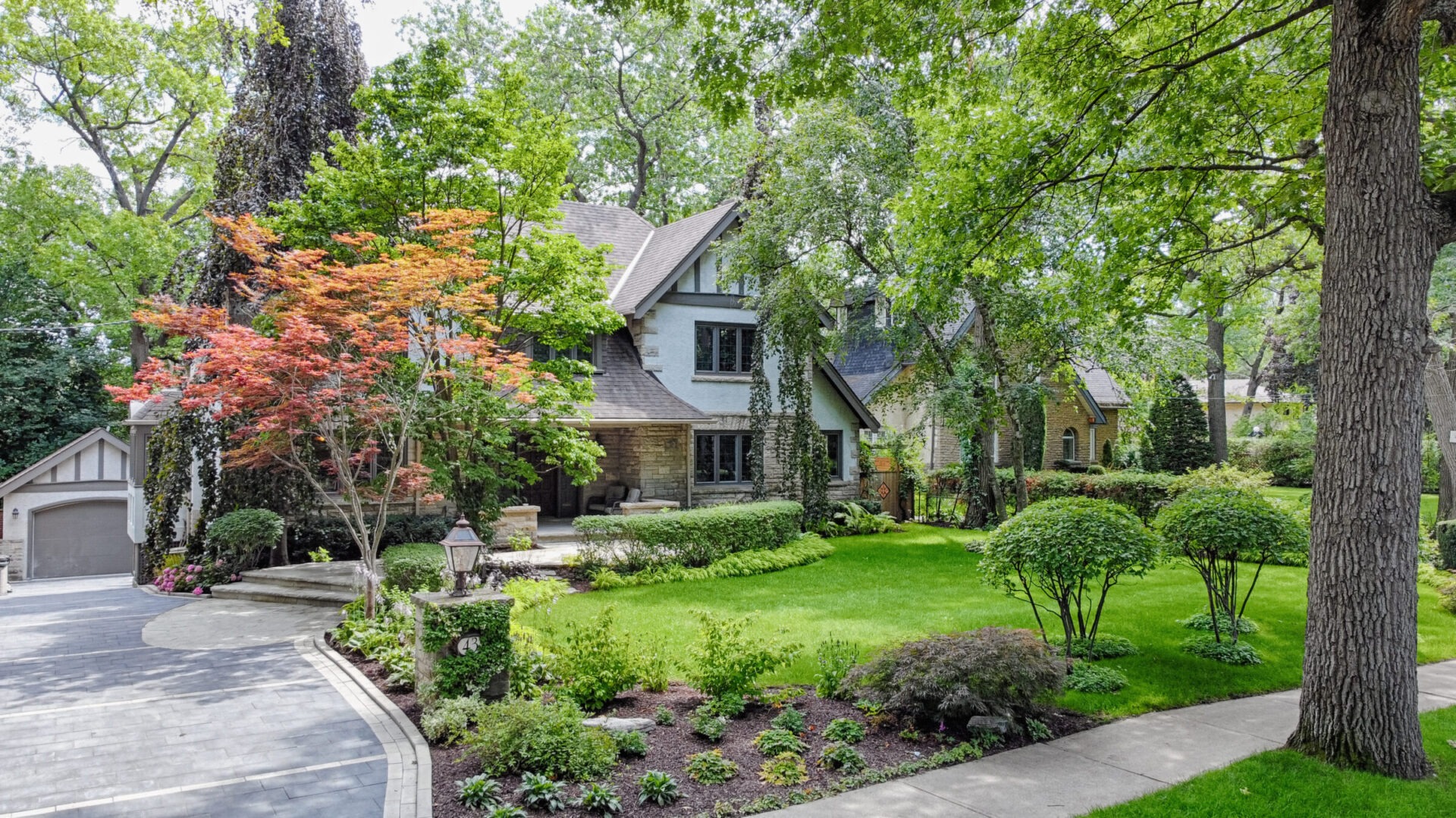 A two-story home with a stone facade is surrounded by lush greenery, manicured bushes, and a red-leaved tree, with a curved driveway leading to a garage.