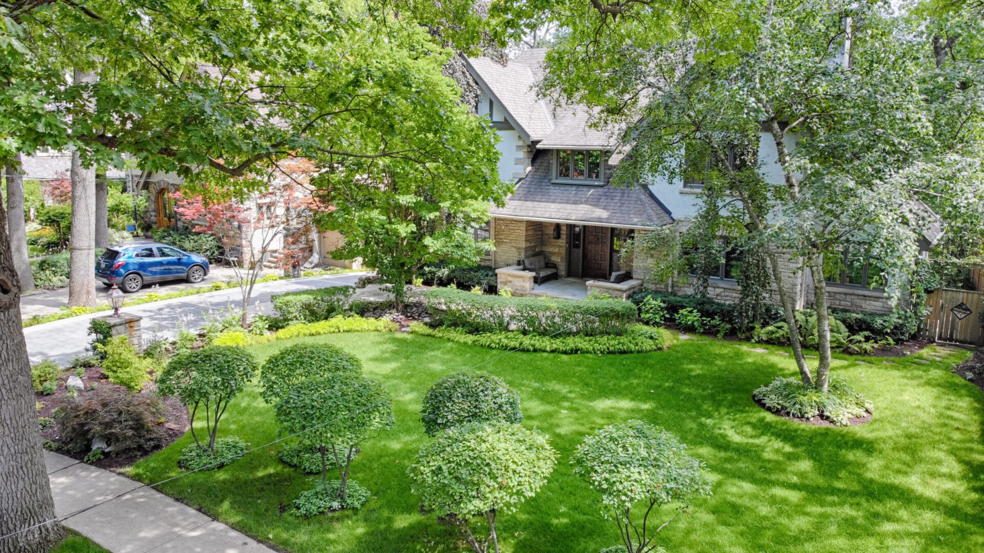 An aerial view of a well-manicured yard with round topiary bushes, a stone house with dark trim, a car parked on a driveway, and lush trees.