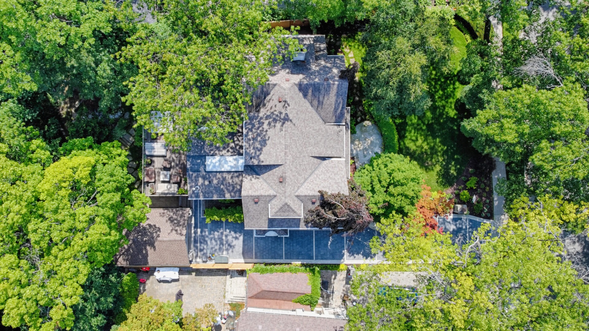 Aerial view of a large, shingled-roof house surrounded by lush greenery with a driveway, white car, and shadow patterns from the surrounding trees.