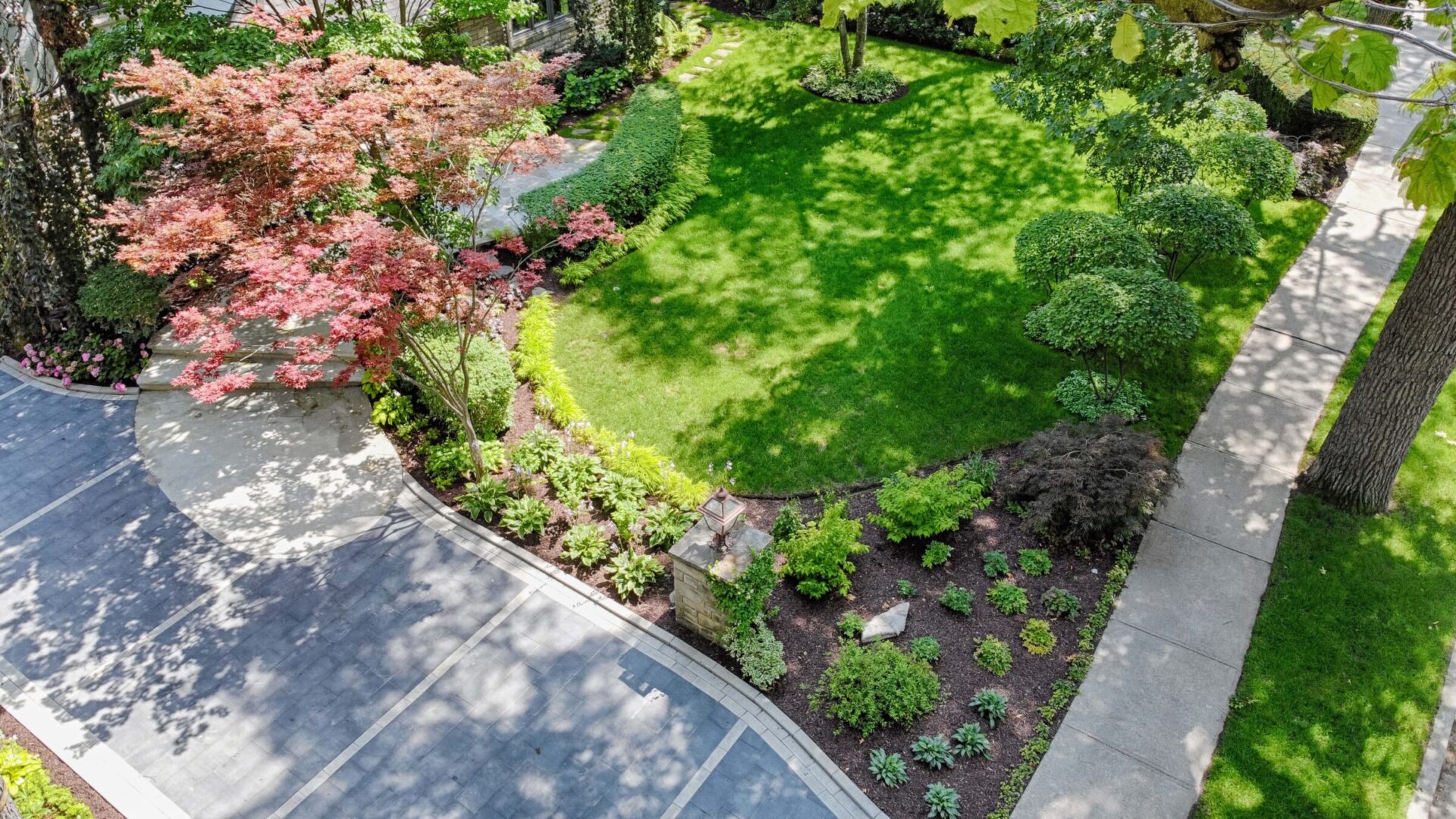 An aerial view of a landscaped garden with a lush green lawn, colorful foliage, a curving pathway, and neatly trimmed bushes on a sunny day.