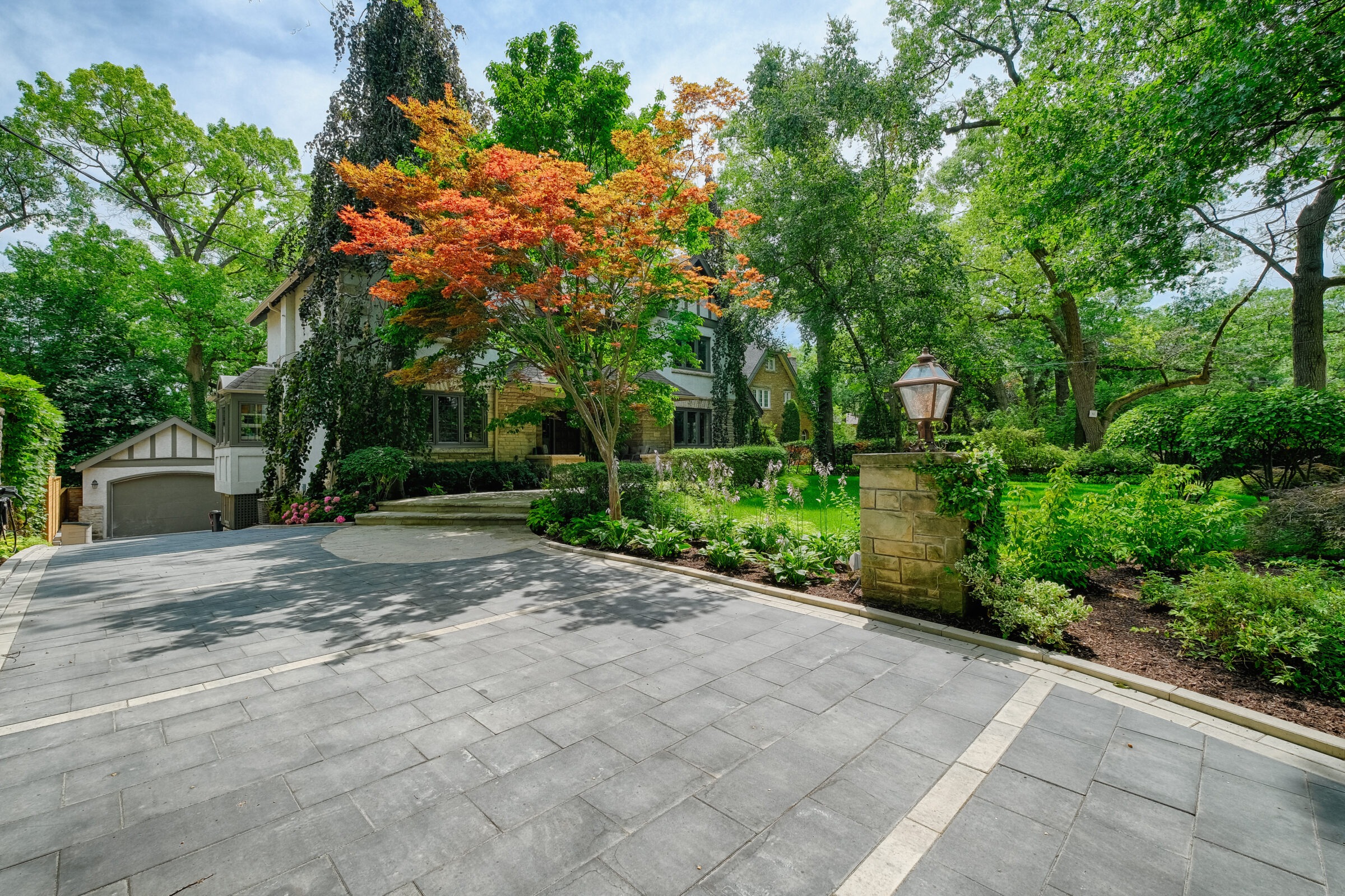 This image shows a large, lush garden with a variety of trees surrounding an elegant stone house. A paved driveway leads to a garage.