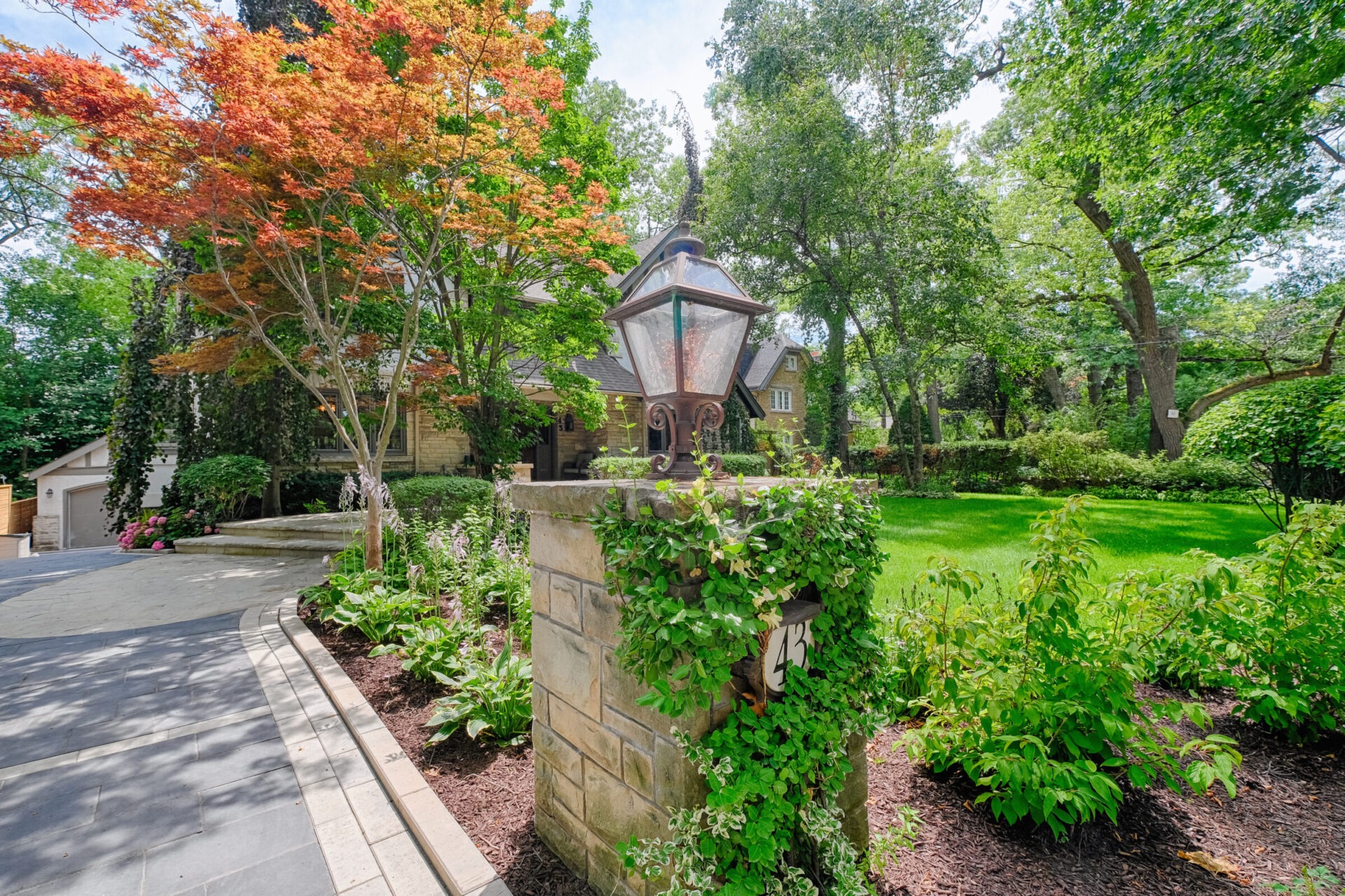 An ornate outdoor lamp post stands atop a stone column amidst lush greenery and gardening, with a curved pathway leading towards a residential backdrop.