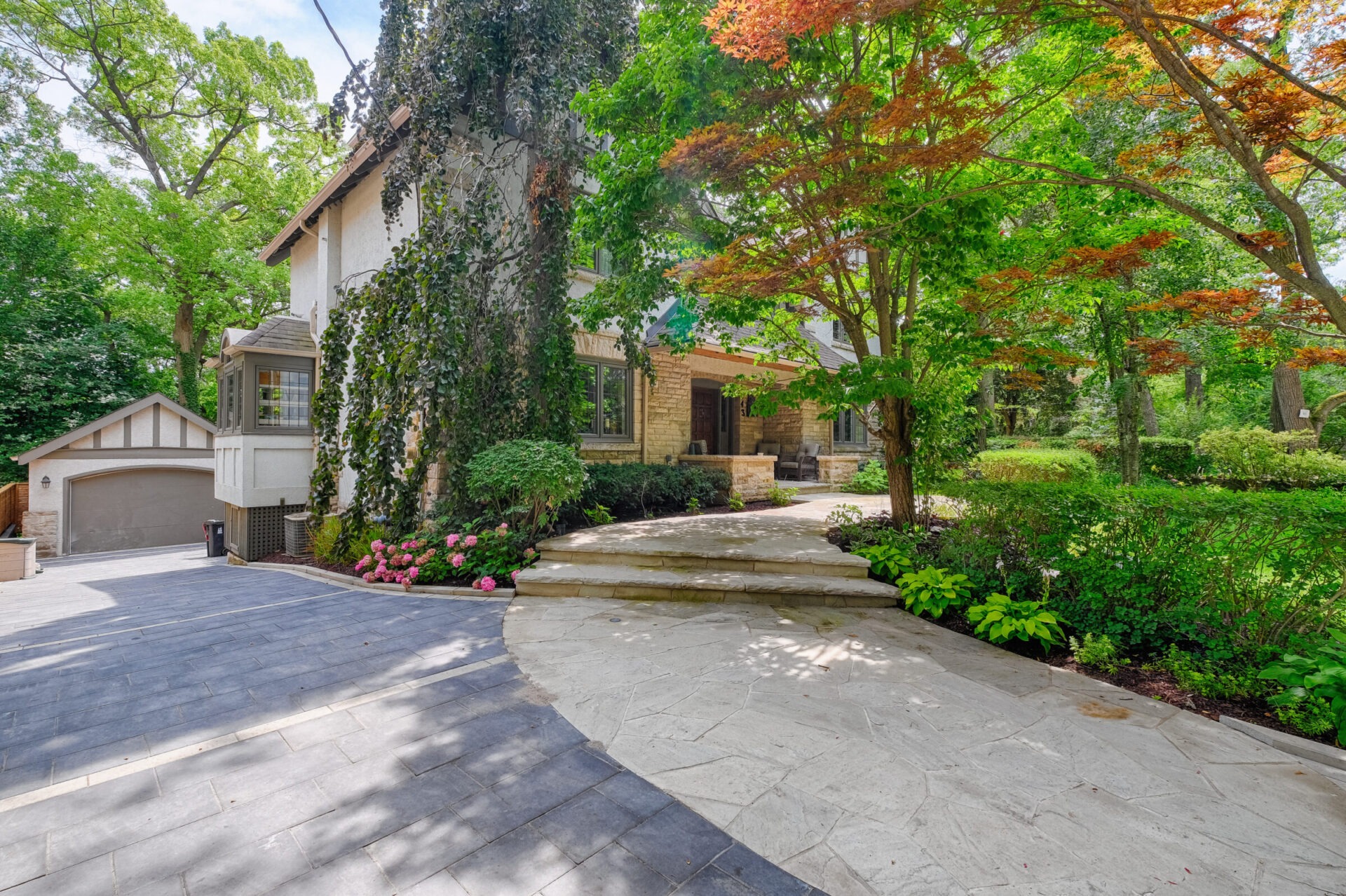 An elegant house with stone facade, surrounded by lush trees and flowering shrubs. A driveway leads to a garage beside a vine-covered wall.