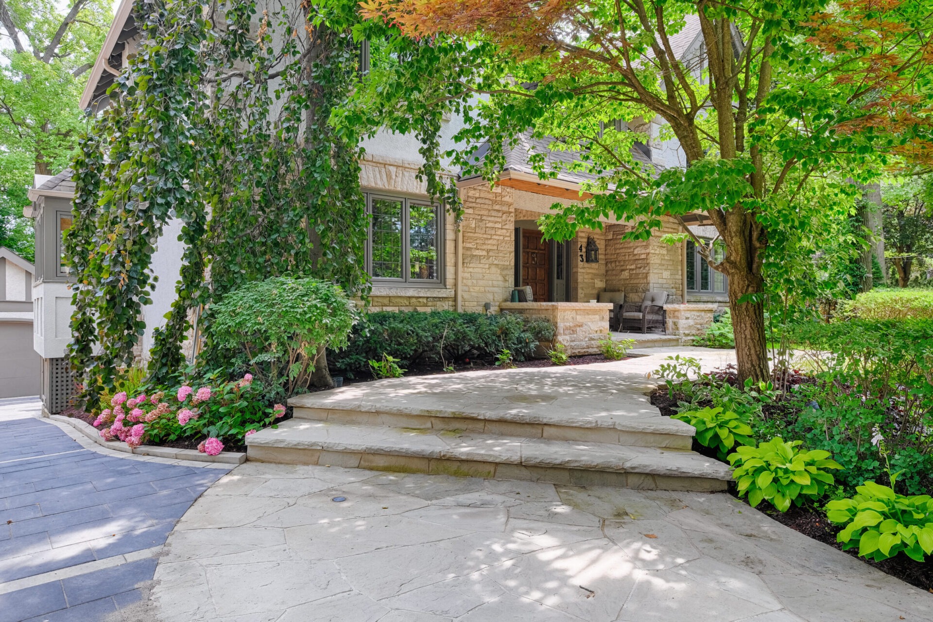 A well-manicured front yard of a two-story house with stone facade, ivy growth, hydrangeas, and a flagstone walkway leading to a cozy porch.