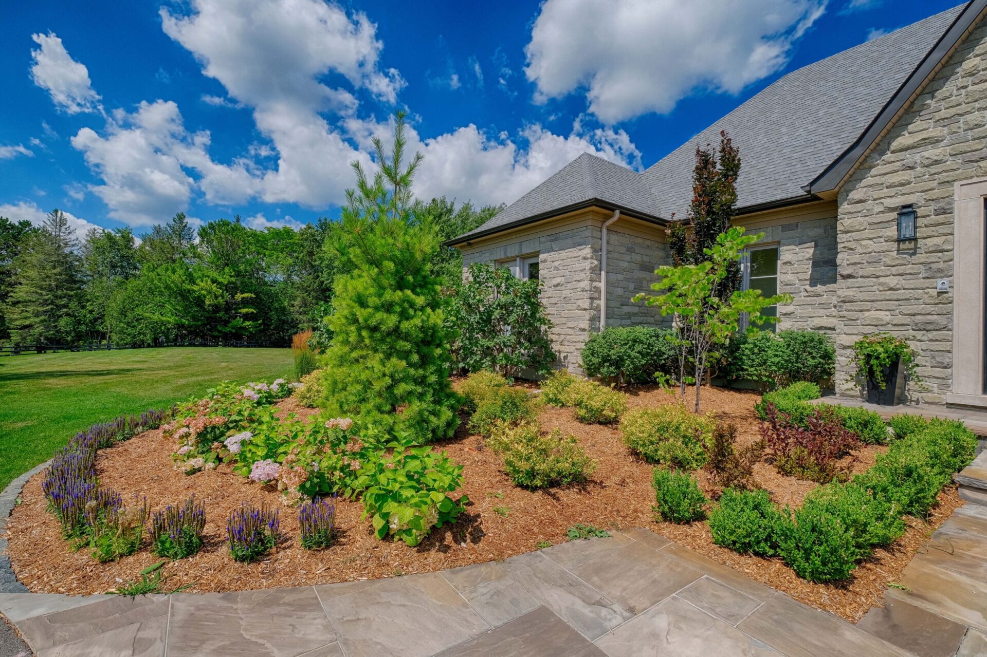 A well-manicured garden with flowering shrubs and a stone house under a sunny sky with fluffy clouds. Green lawn and trees in the background.