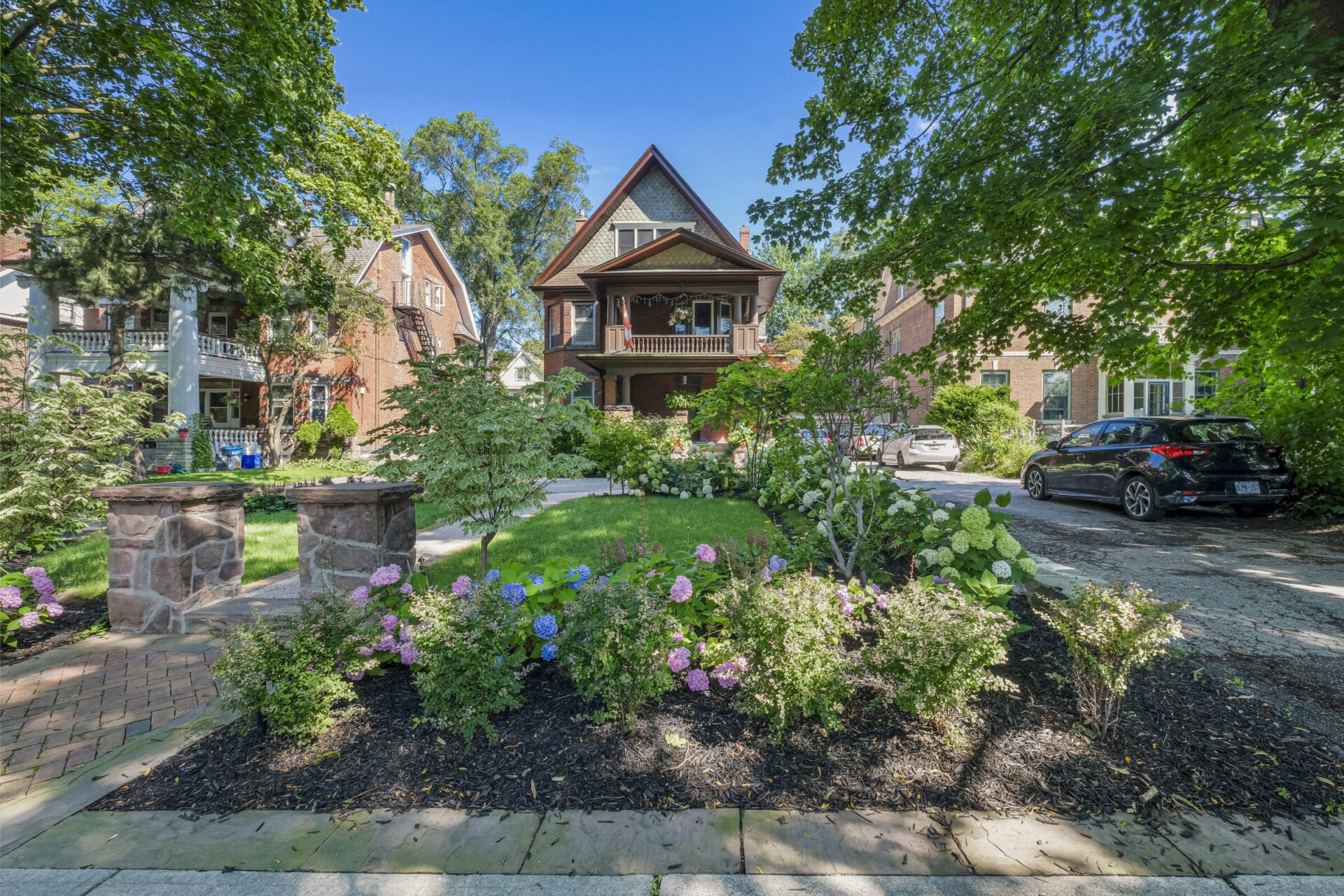 A leafy suburban street with a two-story house featuring a prominent gable and a large covered front porch. Luxuriant gardens and brick walkways enhance the tranquility.