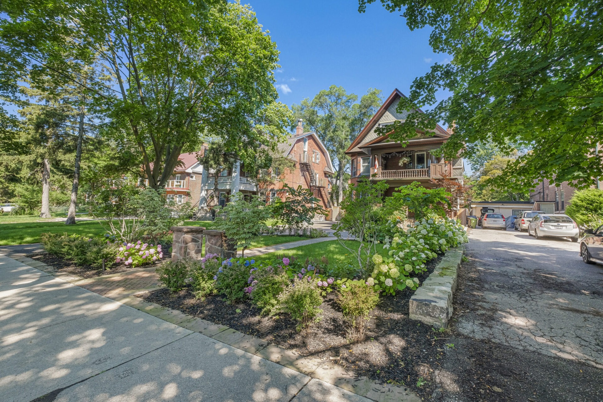 The image shows a serene residential street with lush greenery, multiple houses, a well-manicured garden, and parked cars on a sunny day.