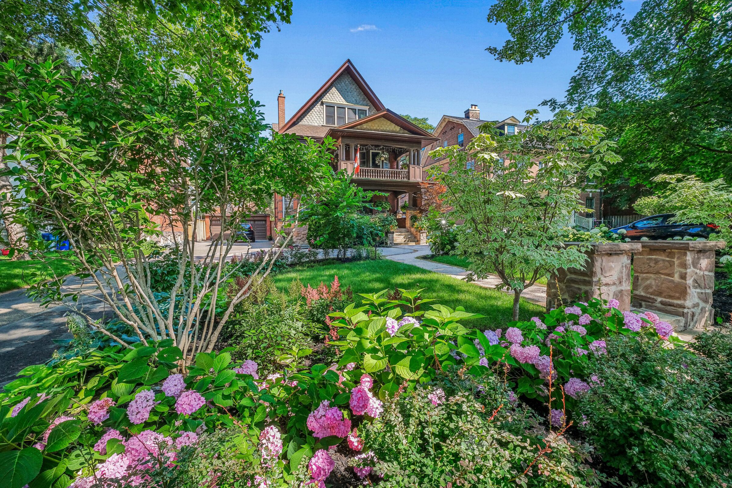 A lush garden with blooming hydrangeas leads to an elegant house with a balcony, shingled roof, and a stone walkway on a sunny day.