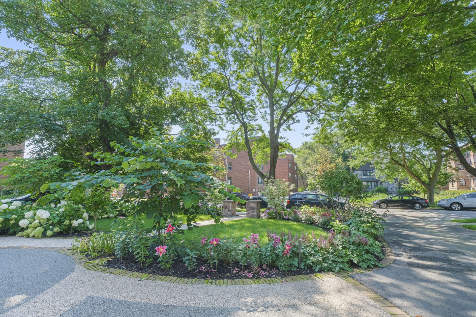 This image portrays a tranquil residential street with lush greenery, flowering plants, parked cars, and brick buildings under a clear blue sky.