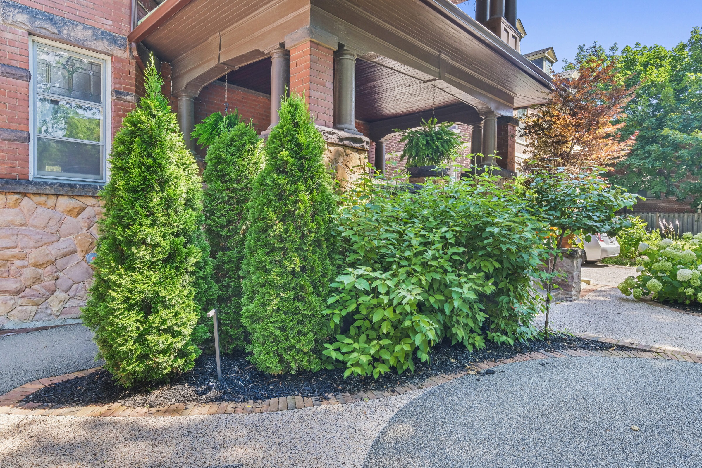 A brick house with a wooden porch has neatly landscaped green shrubs and trees in a residential setting with a clear blue sky overhead.