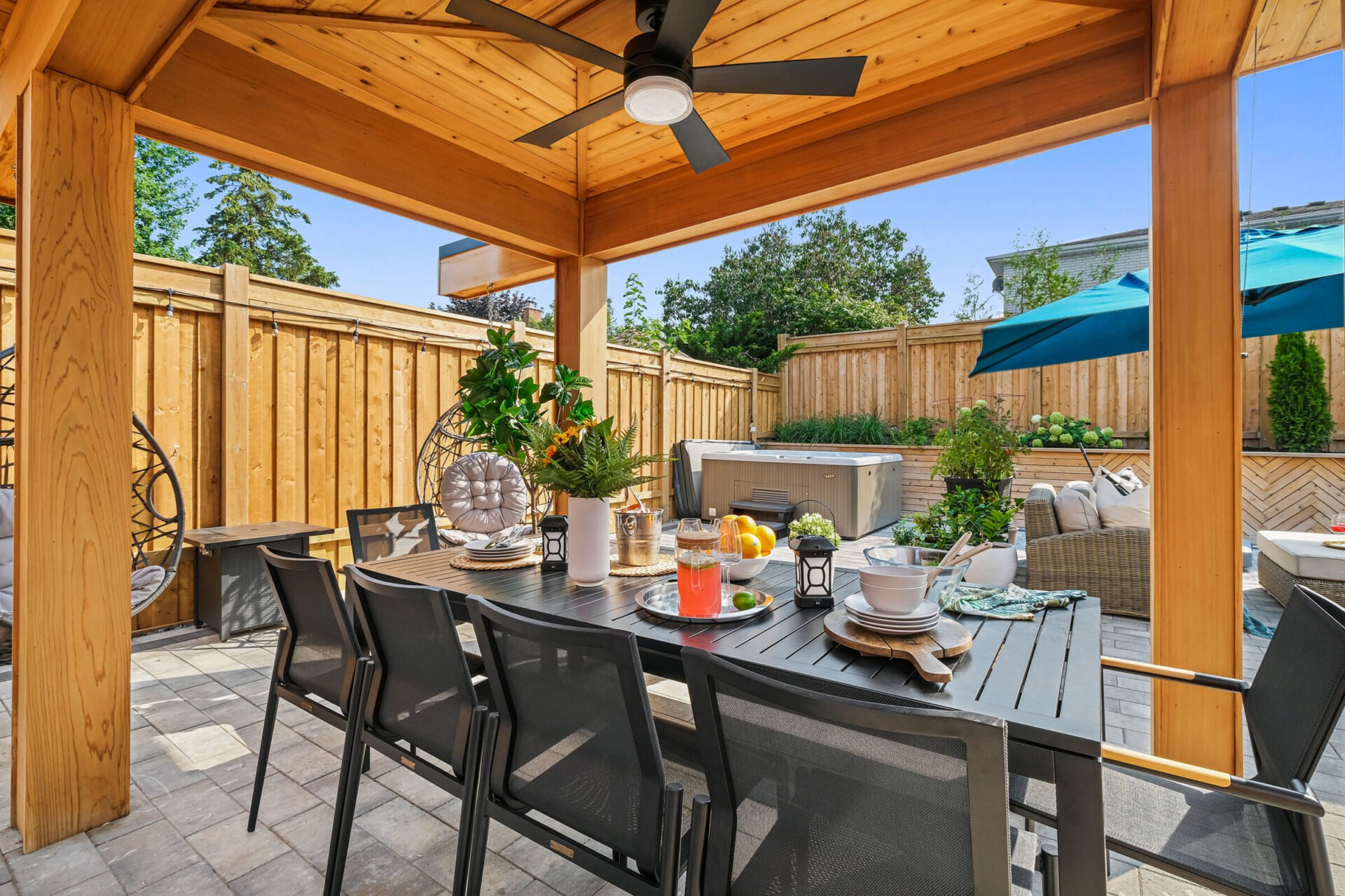 This outdoor patio features a wooden dining set, ceiling fan, hot tub, and comfortable seating area, surrounded by a privacy fence under clear skies.