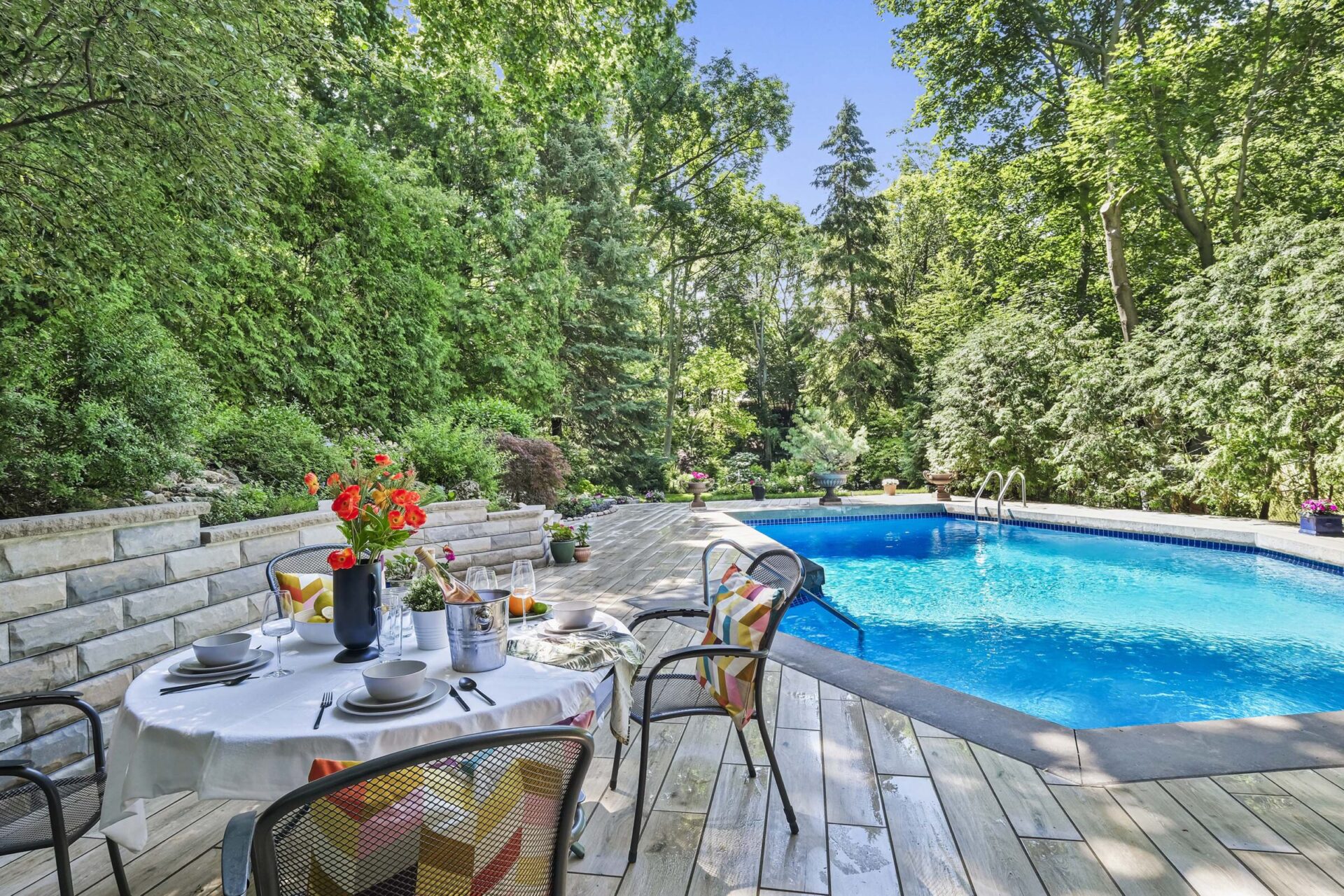 A serene outdoor setting featuring a dining table with a vase of red flowers; a pool with clear blue water; surrounded by lush green trees.