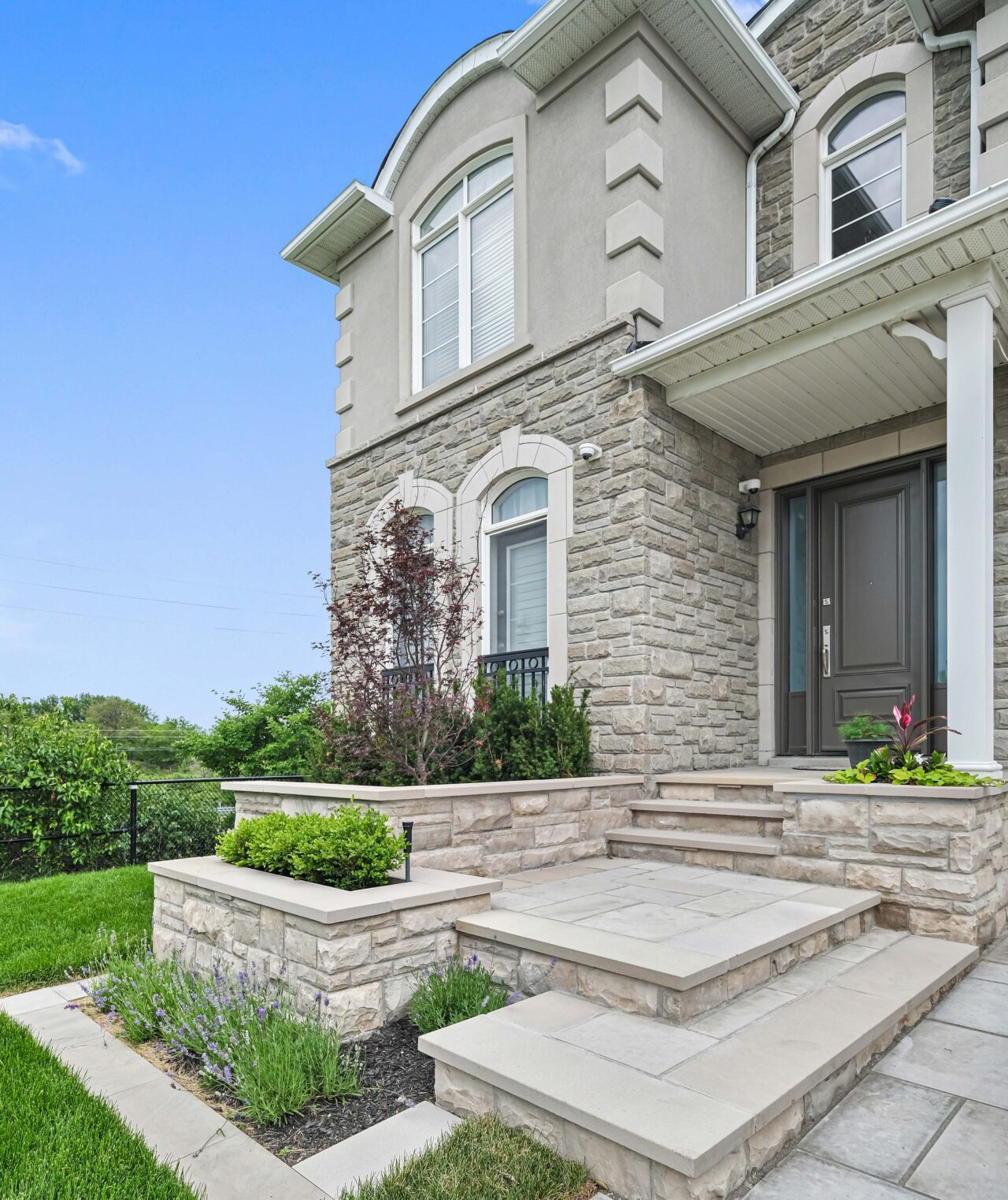 This image shows the exterior of a two-story house with stone and stucco facade, steps leading to a front door, and landscaped gardens under a clear sky.