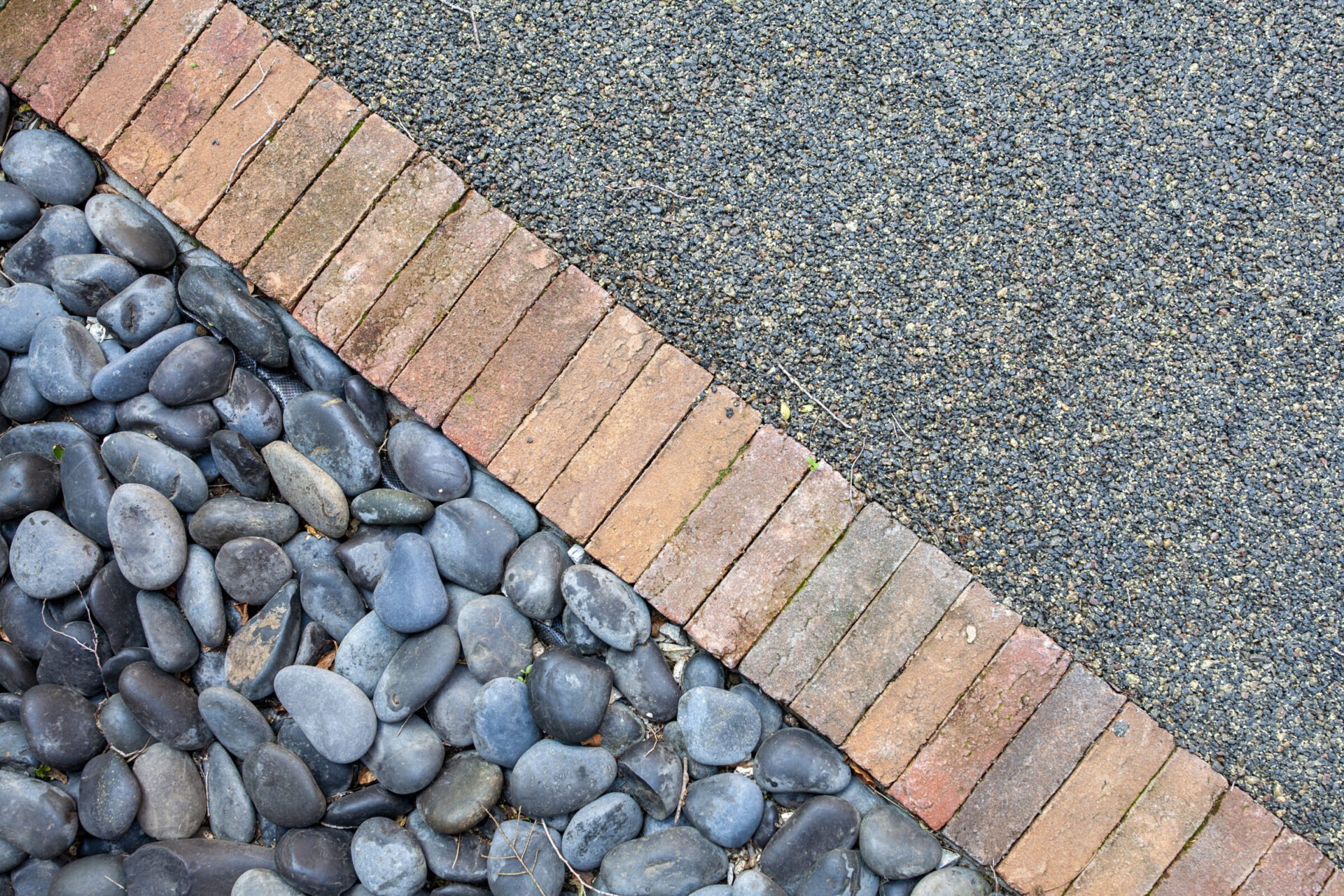 This image shows a textured surface transition with smooth dark pebbles, red brick edging, and a coarse gravel pathway in natural daylight.