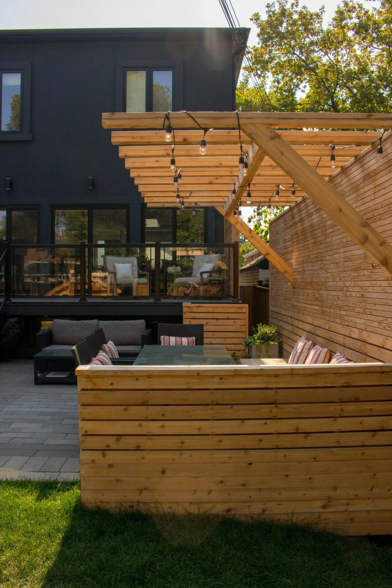 A modern backyard with a pergola, string lights, wooden deck, outdoor furniture, large windows, and a two-story dark-colored house in the background.