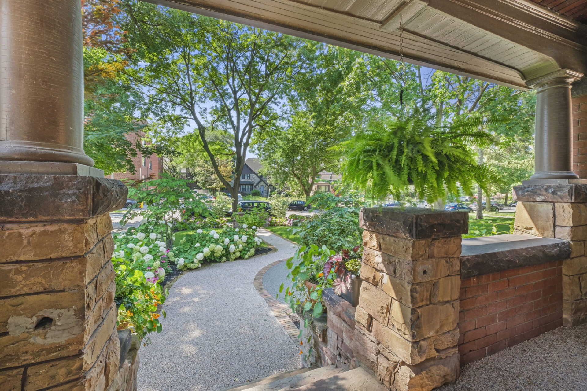 This image shows a verdant garden view from a porch with stone columns, hanging ferns, hydrangeas, and a winding path leading to a residential area.