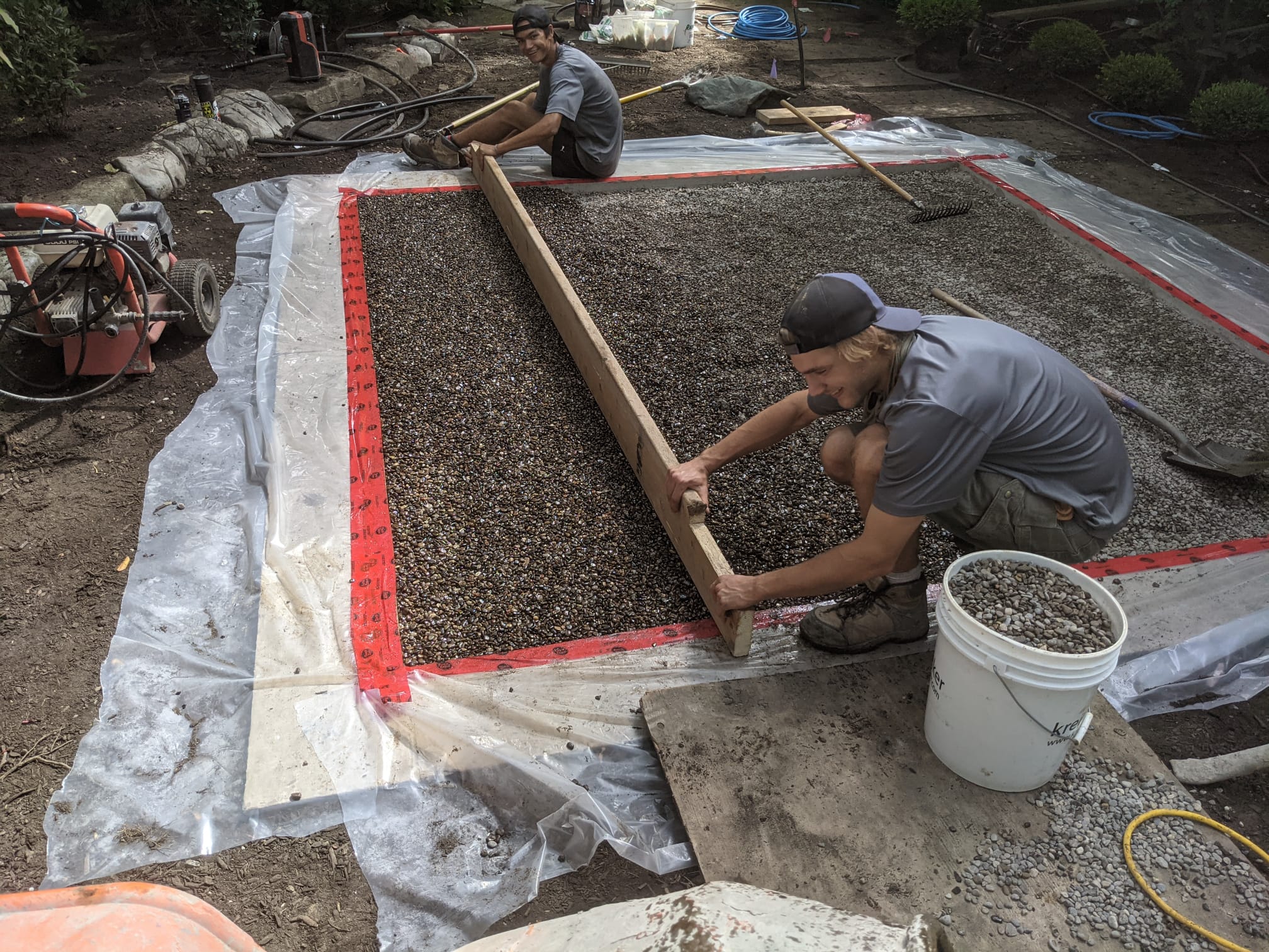 Two people are working on a construction site, leveling gravel within a wooden frame, surrounded by tools and landscaping equipment.