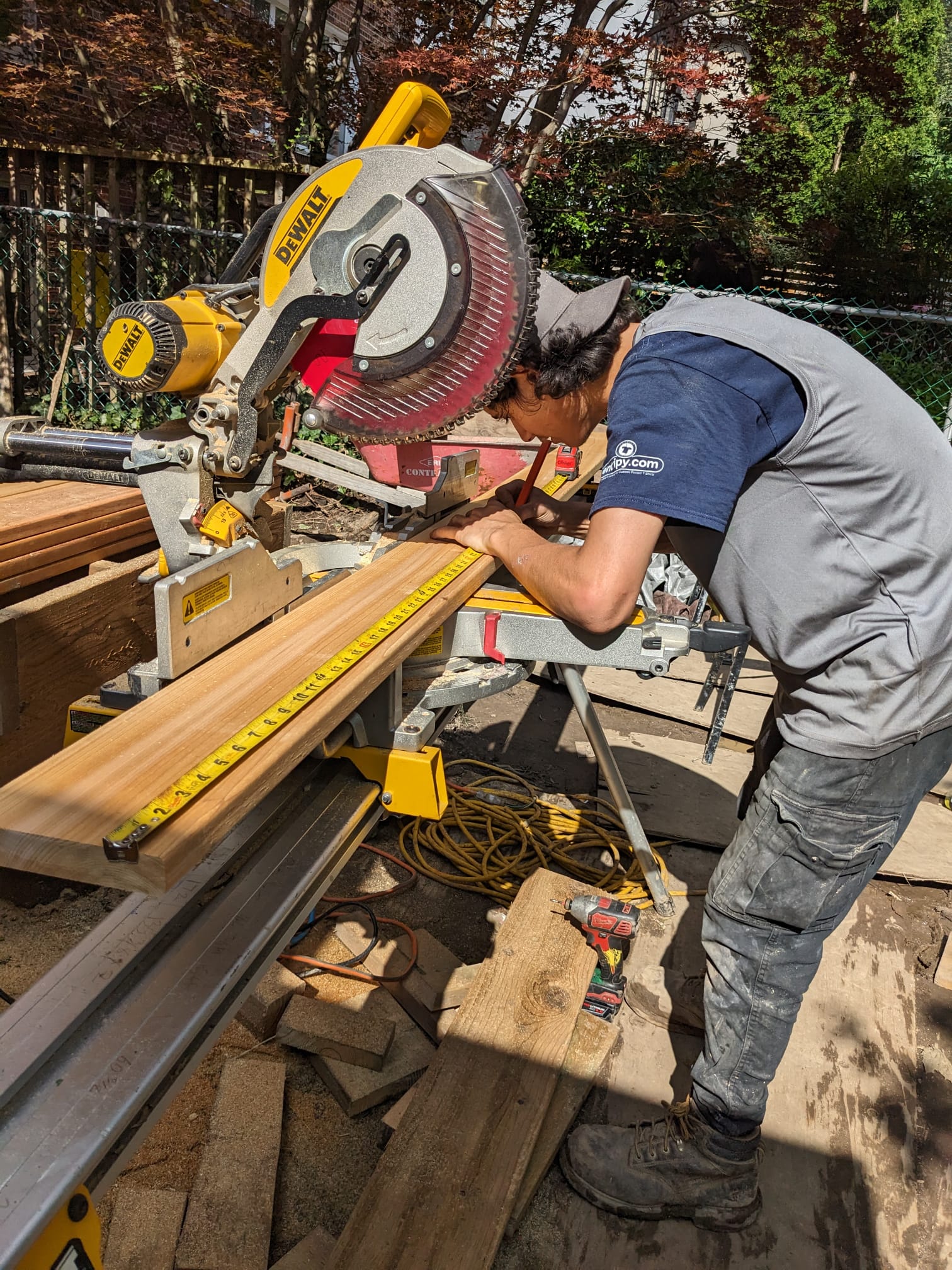 A person is using a miter saw to cut wood outdoors. There's a measuring tape, safety equipment, and construction materials scattered around the workspace.