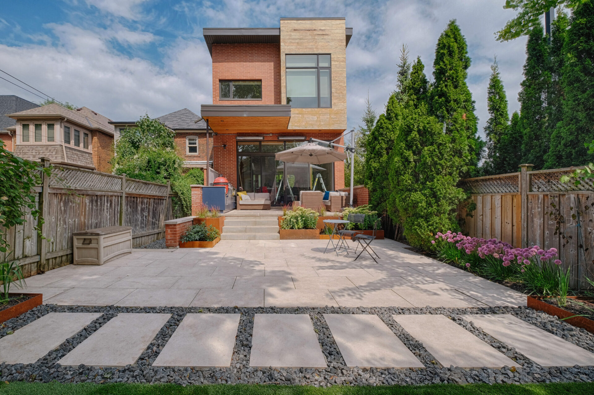 This image shows a well-manicured backyard with a modern two-story house, large patio area, outdoor furniture, greenery, and stepping stones leading to the porch.