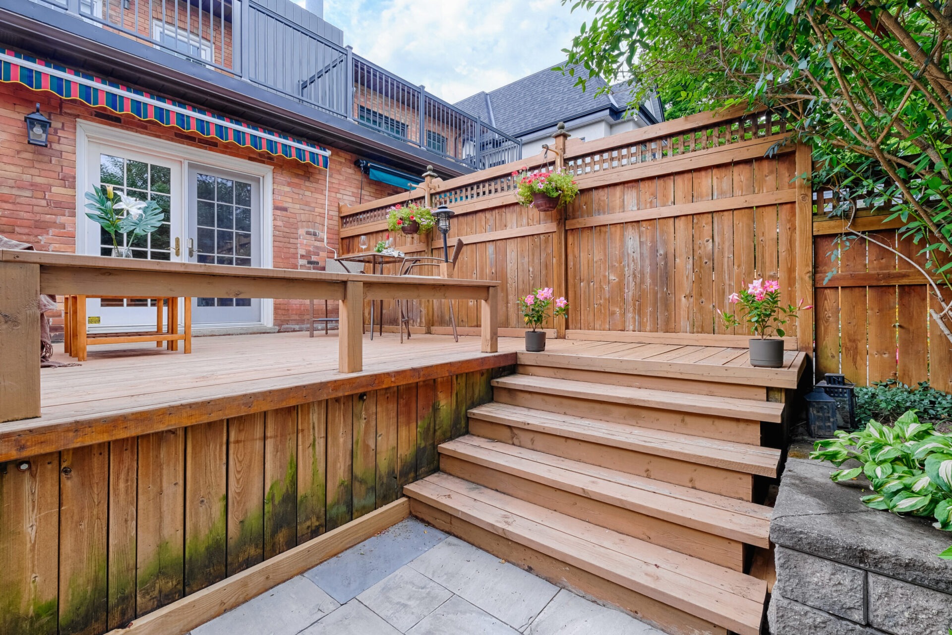 A wooden deck with steps features a table, chairs, potted plants, and a privacy fence. It’s adjoined to a building with a balcony above.