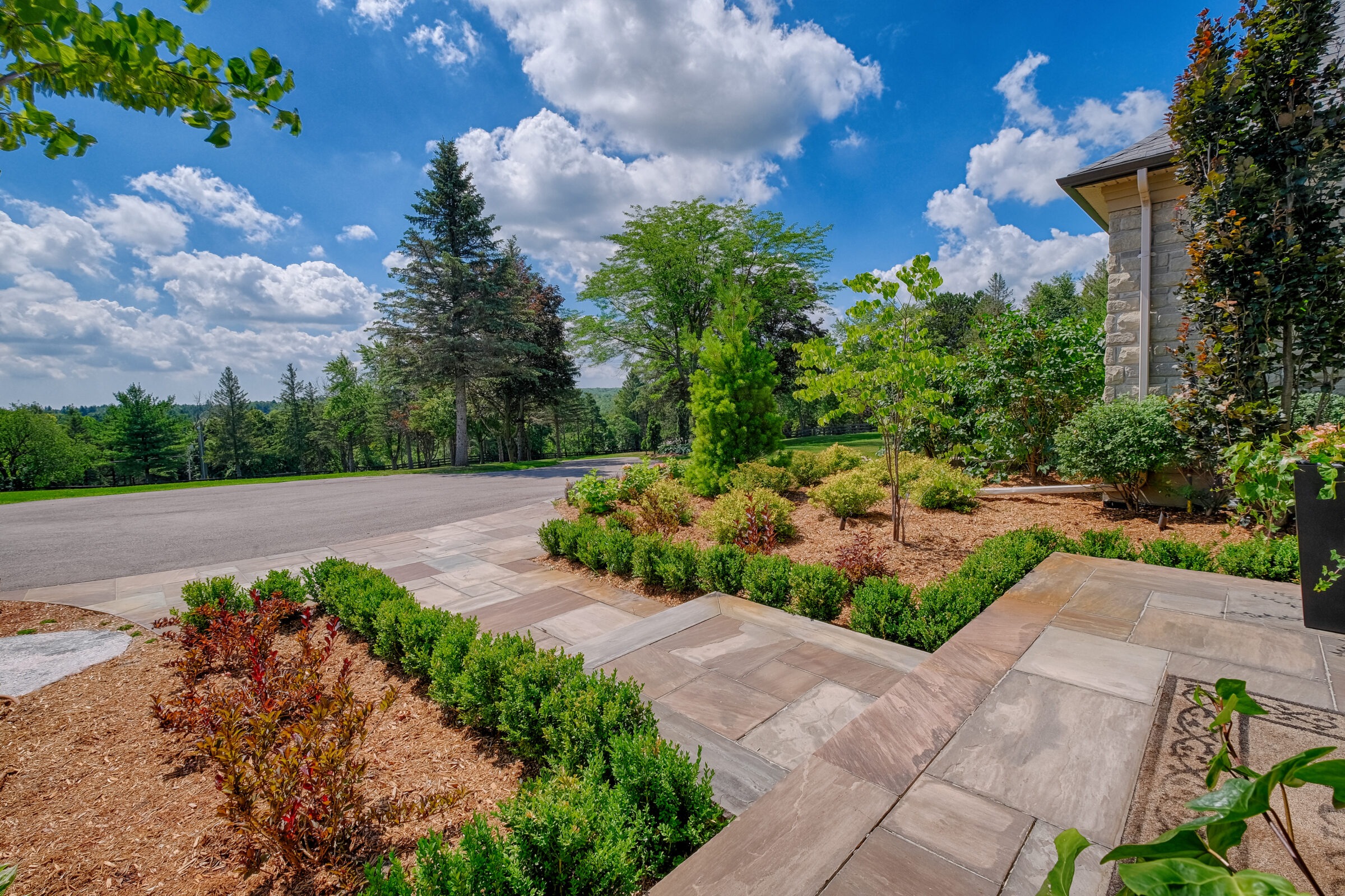 The image shows a landscaped garden with neatly arranged bushes and trees, leading to a stone pathway beside a large house under a sunny sky.