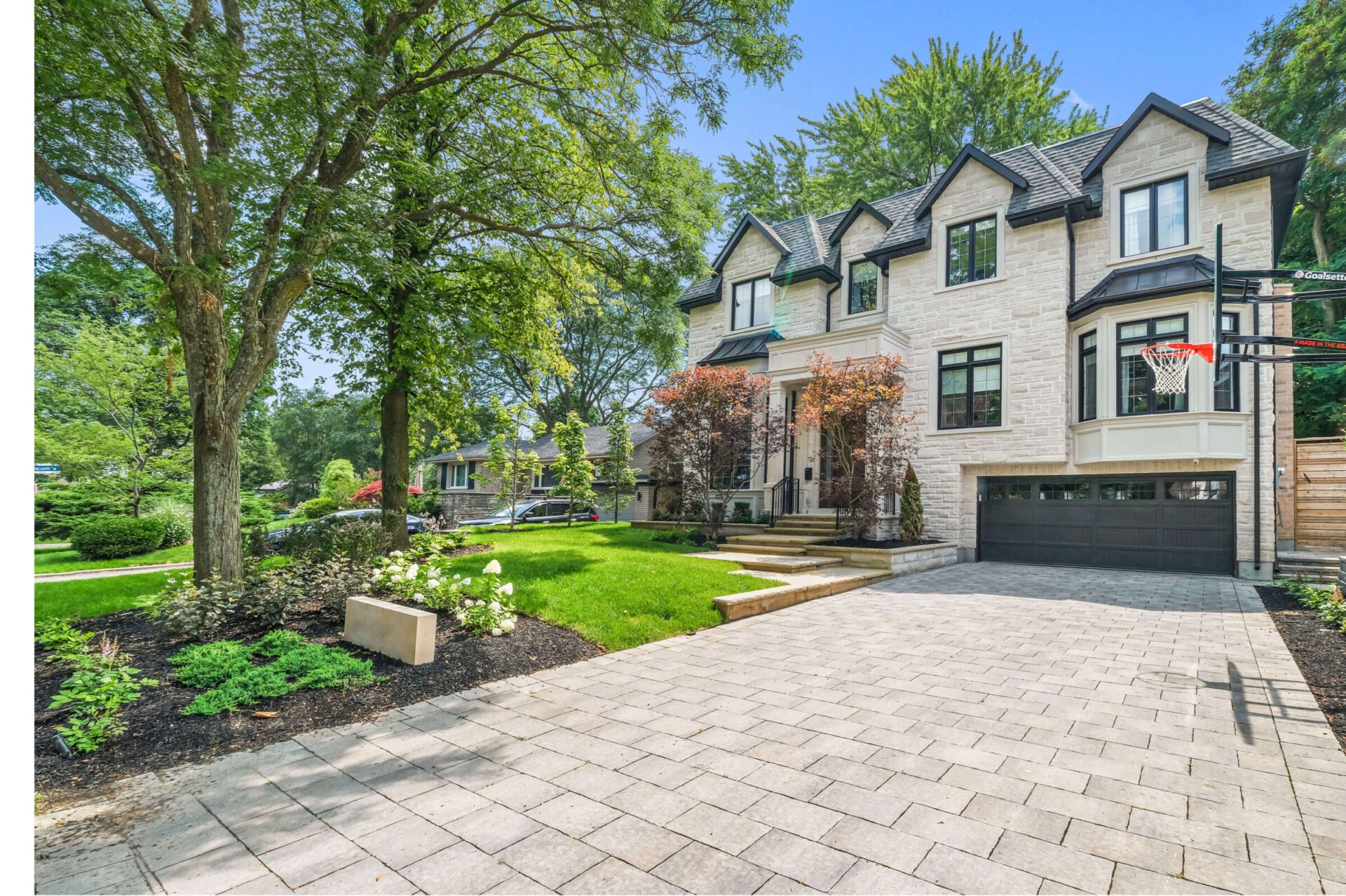 This image shows a two-story residential home with a stone facade, surrounded by trees, landscaped garden, paver driveway, and a basketball hoop.