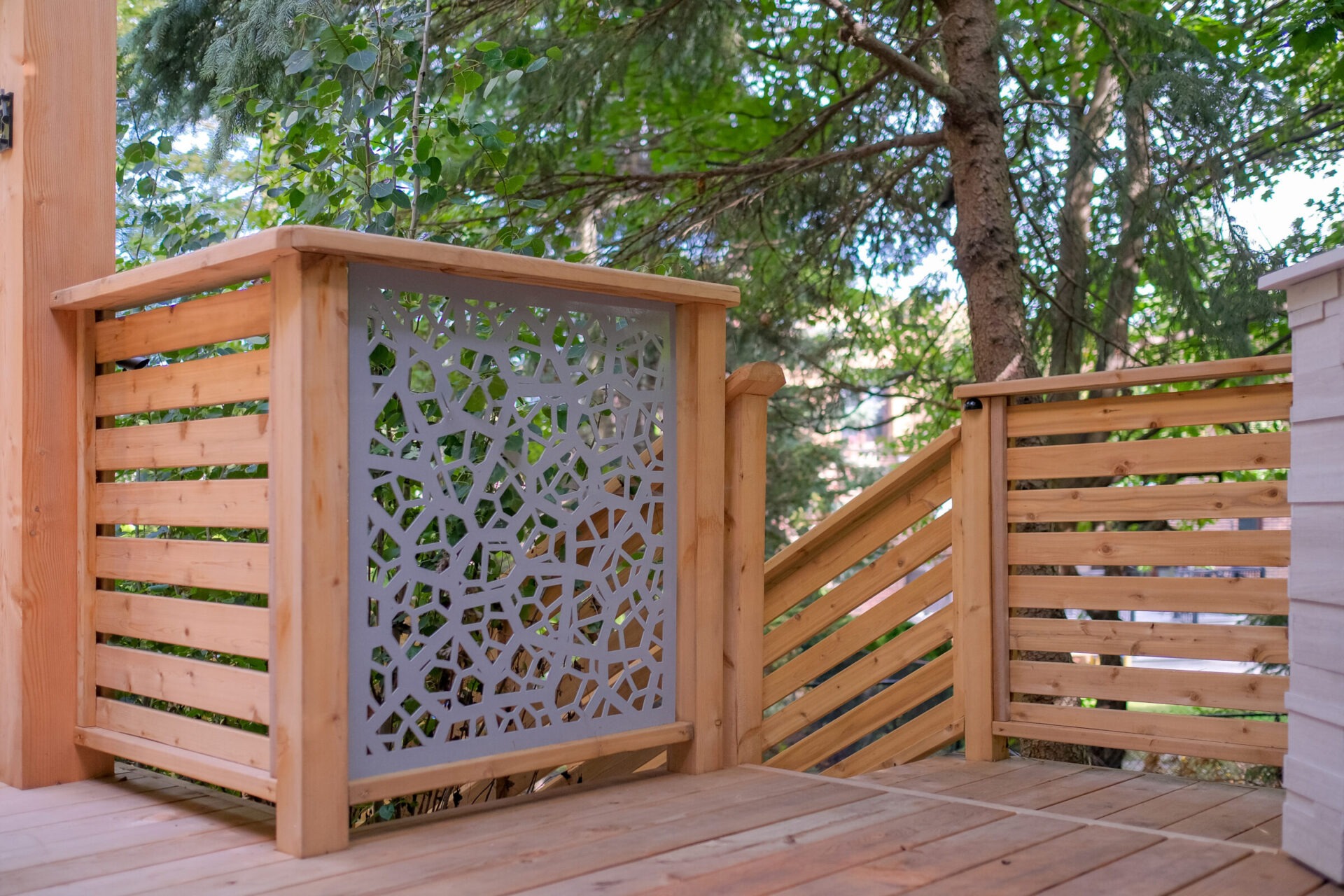 The image shows a wooden deck with a decorative laser-cut panel and horizontal railings, surrounded by green trees in a tranquil outdoor setting.