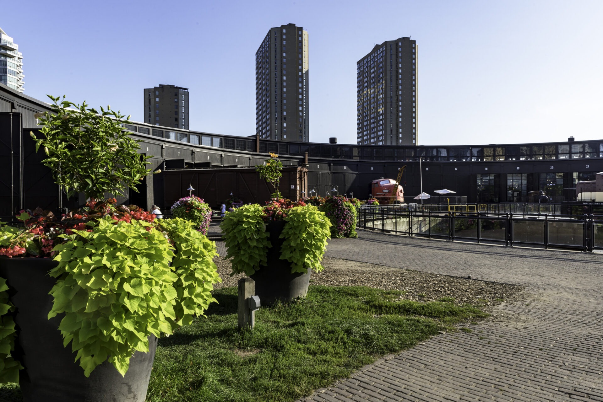 Urban scene with twin high-rise buildings in the background, large planters with lush greenery in the foreground, and a person visible near buildings.