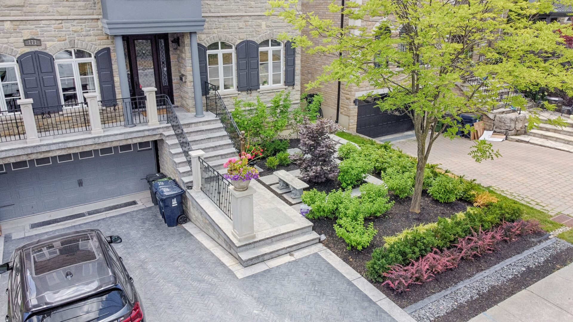 Aerial view of a neatly landscaped residential property with stone facade, a grey garage door, a black SUV, and green shrubbery surrounding a tree.