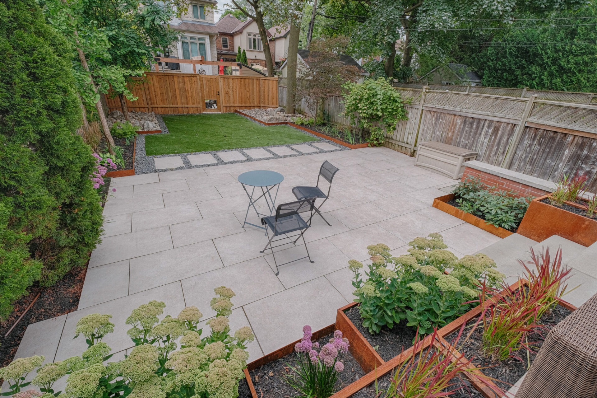 This image shows a neatly landscaped backyard with a patio area, green lawn, plants, outdoor furniture, and a wooden fence surrounding the space.