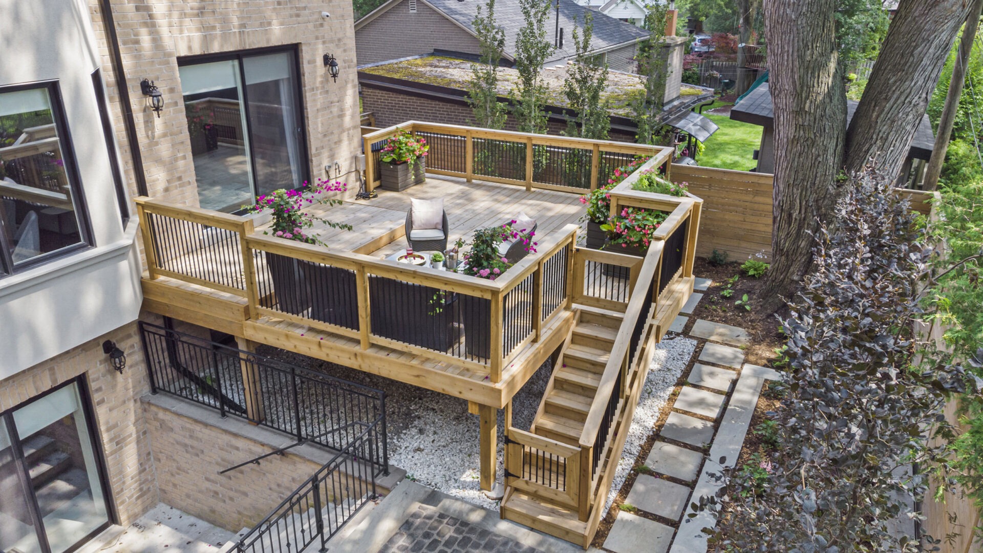 Aerial view of a residential backyard with a wooden deck, planter boxes with flowers, outdoor furniture, pathway, and trees surrounding the area.