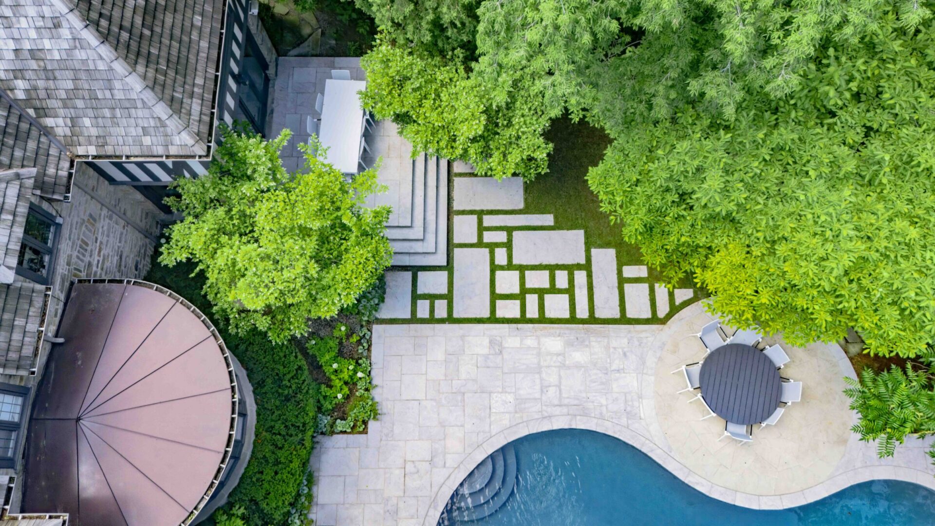 Aerial view of a landscaped yard with a pool, stepping stones, lush trees, a patio umbrella, and an outdoor seating area adjacent to a building.