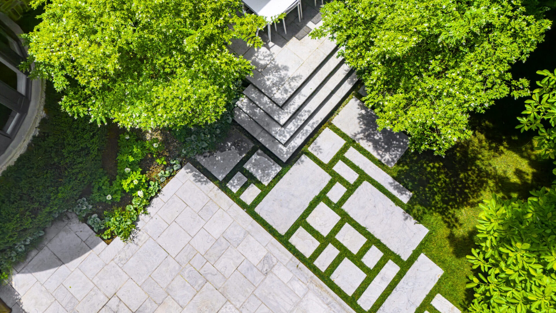 This image shows an aerial view of a symmetrically designed garden with patterned paving, lush greenery, trees, and a section with outdoor furniture.