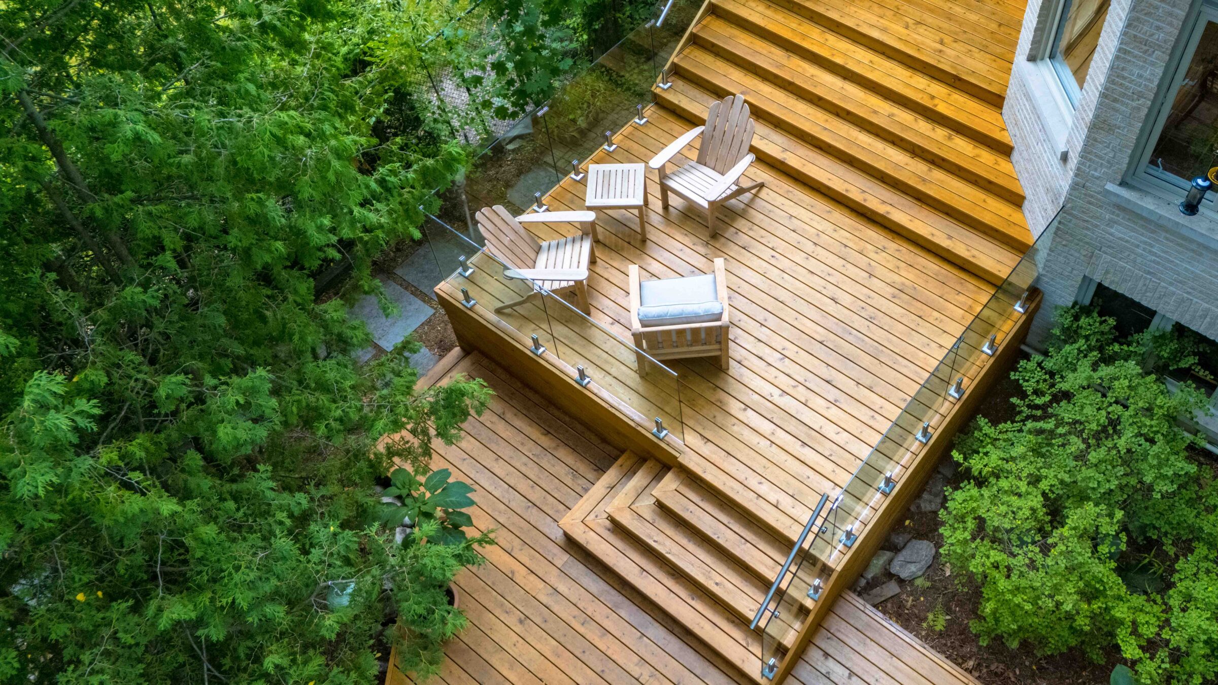 Aerial view of a wooden deck with two Adirondack chairs and a table, surrounded by lush greenery next to a modern house.