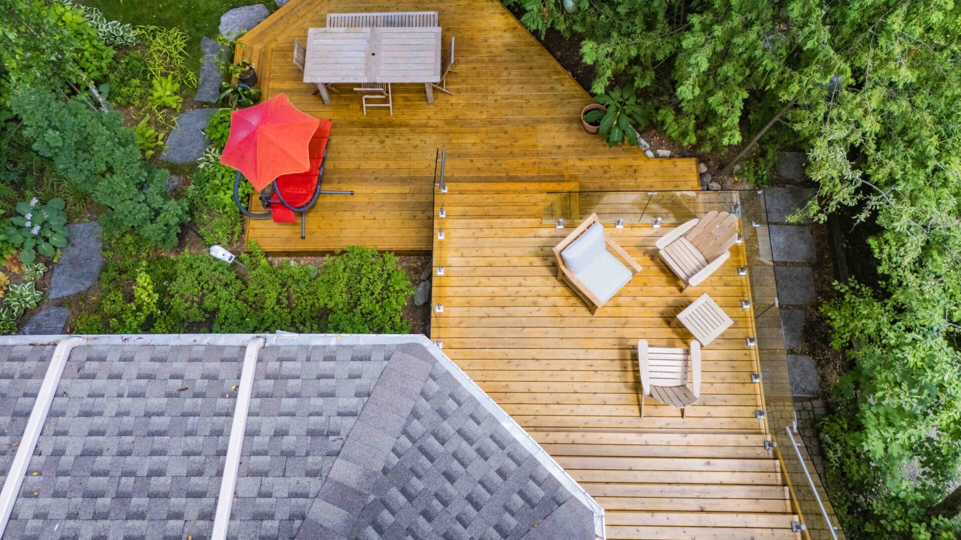 An aerial view shows a wooden deck with outdoor furniture bordered by lush greenery and part of a roof with shingles in the foreground.