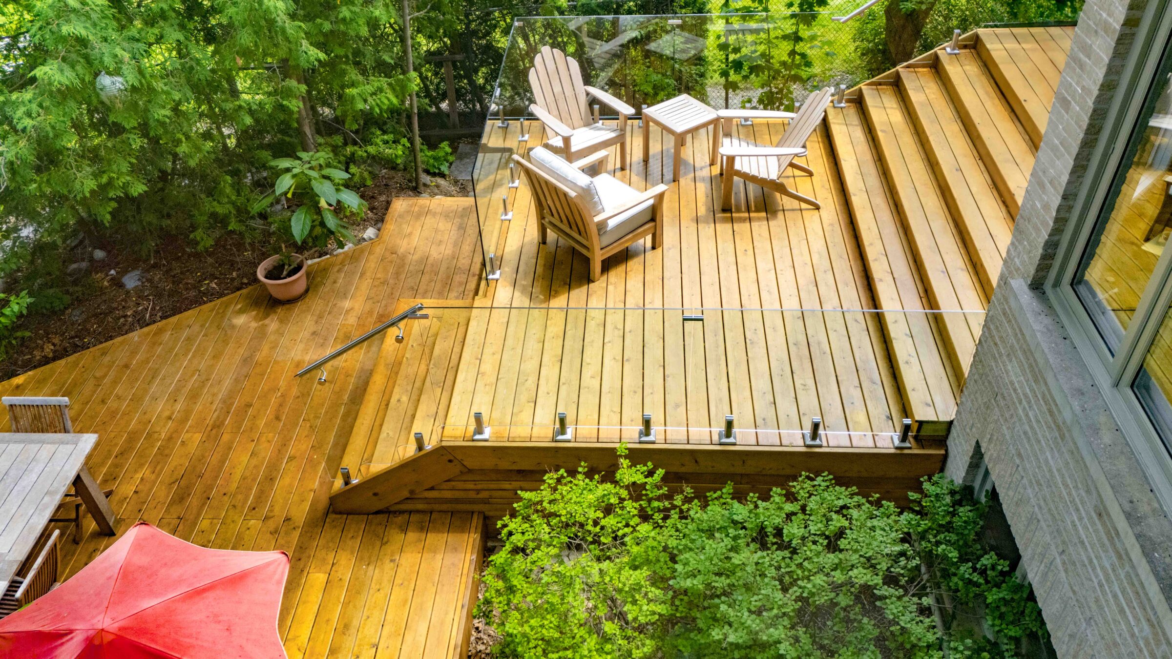 A wooden deck with modern outdoor furniture surrounded by lush greenery, featuring a glass railing, under a clear sky, attached to a stone house.
