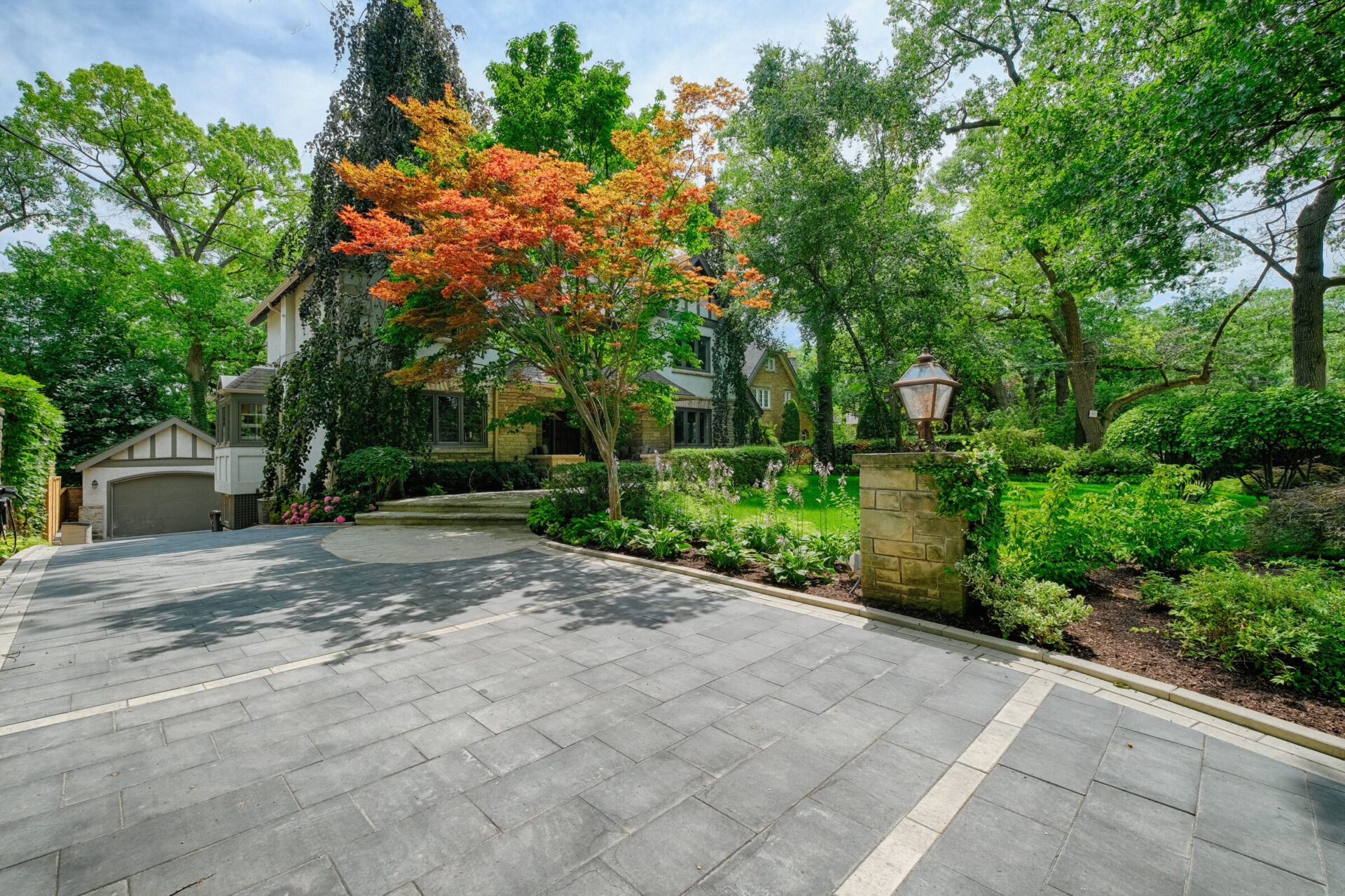 An elegant house with a stone facade sits among lush greenery, highlighted by a vibrant red-orange tree, with a smooth driveway leading towards a garage.