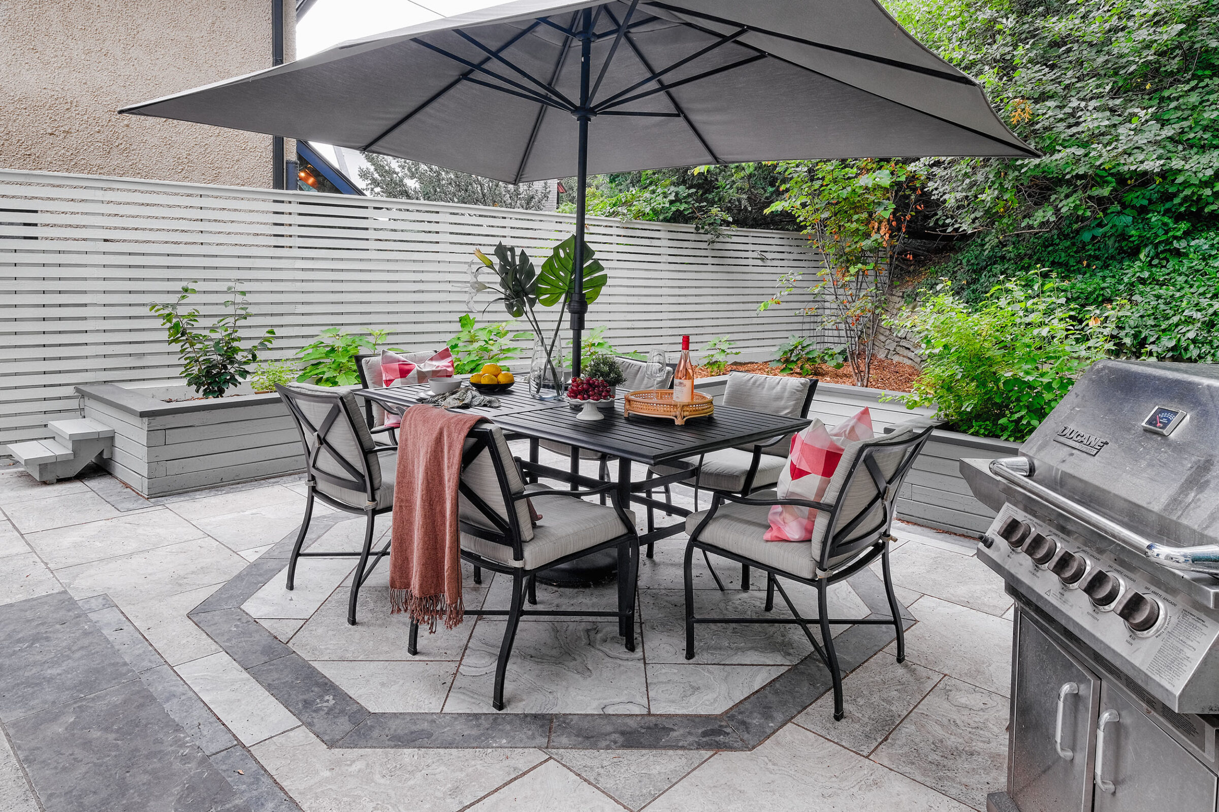 A cozy outdoor patio with a large umbrella, dining table set, cushions, barbecue grill, and surrounded by a wooden fence and greenery.