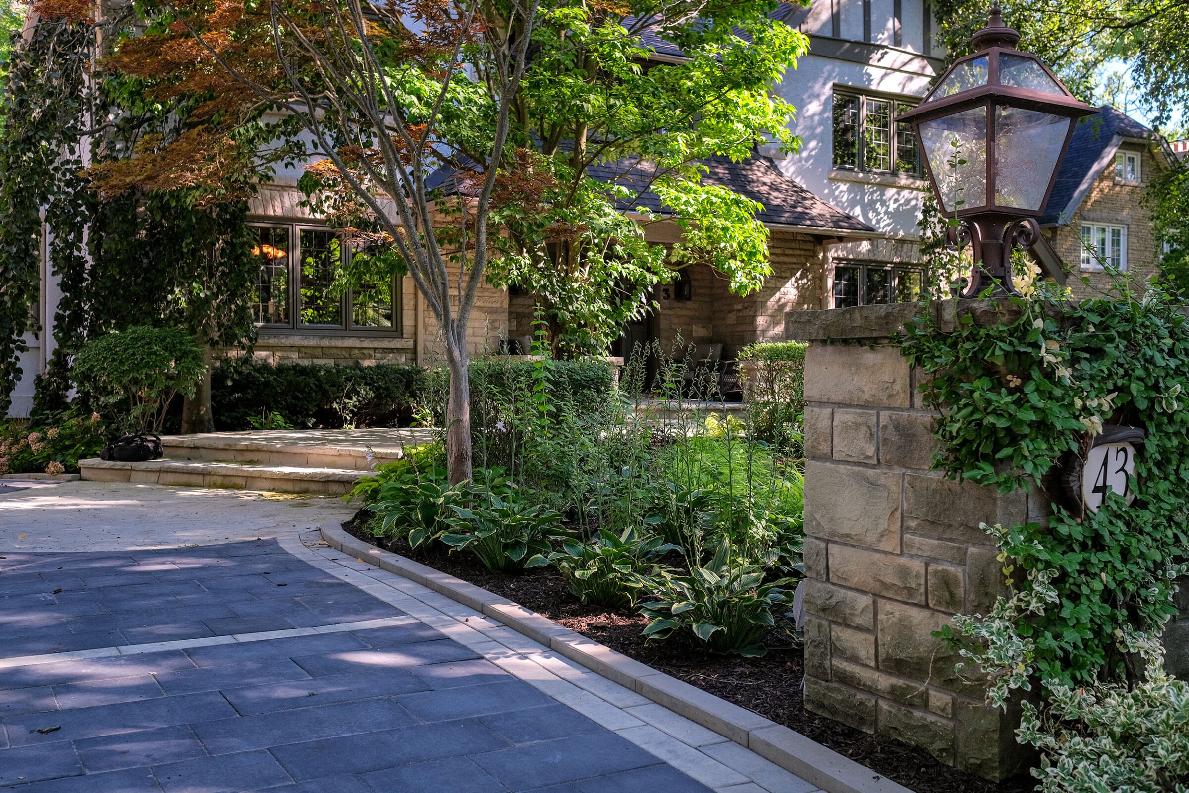 This image showcases an elegant house with stone steps, lush garden, a stately lamp post, and a clear blue sky peeking through the foliage.