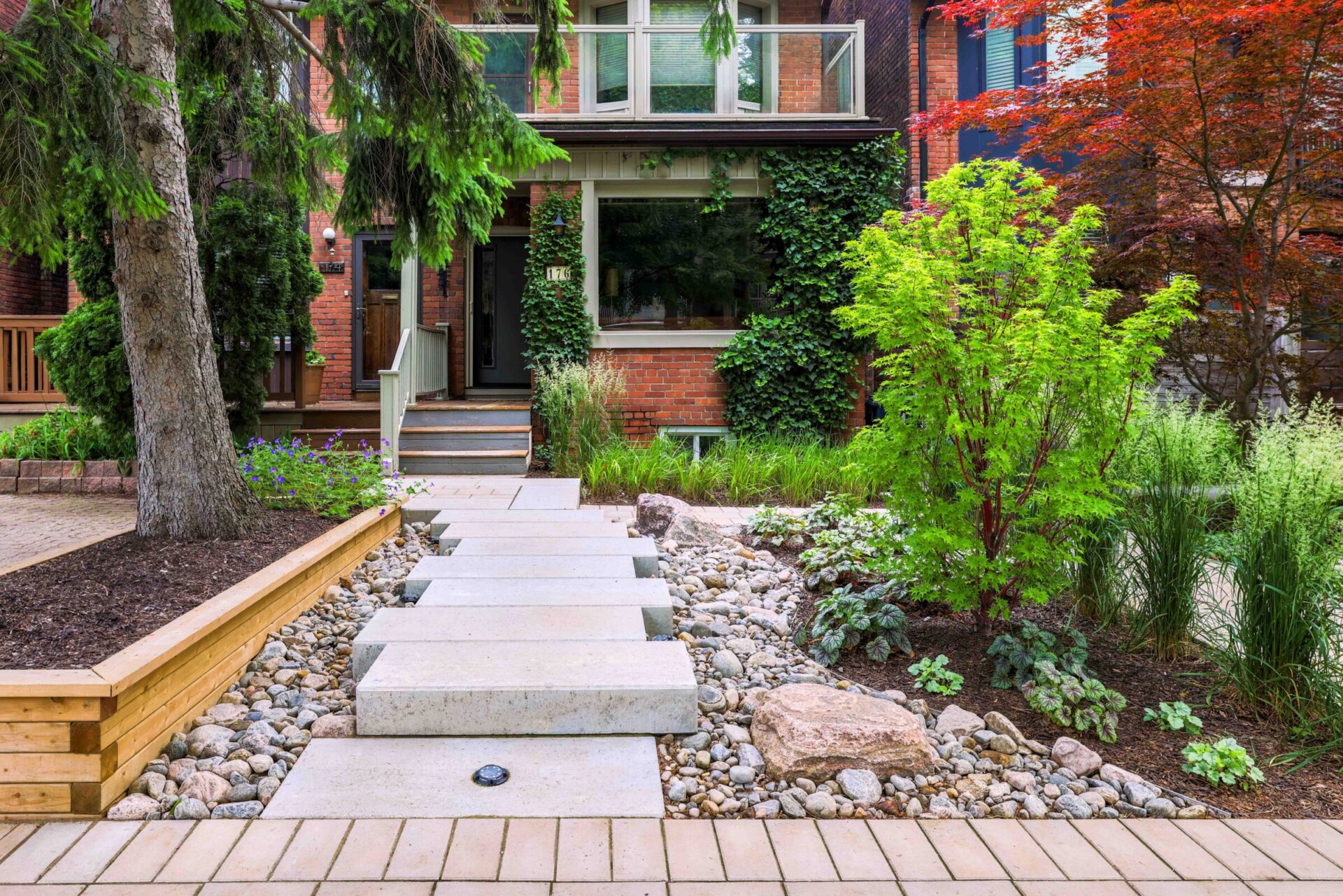 A picturesque residential front yard with lush greenery, a tree, a stone pathway, and a red-brick house with a wooden front porch.