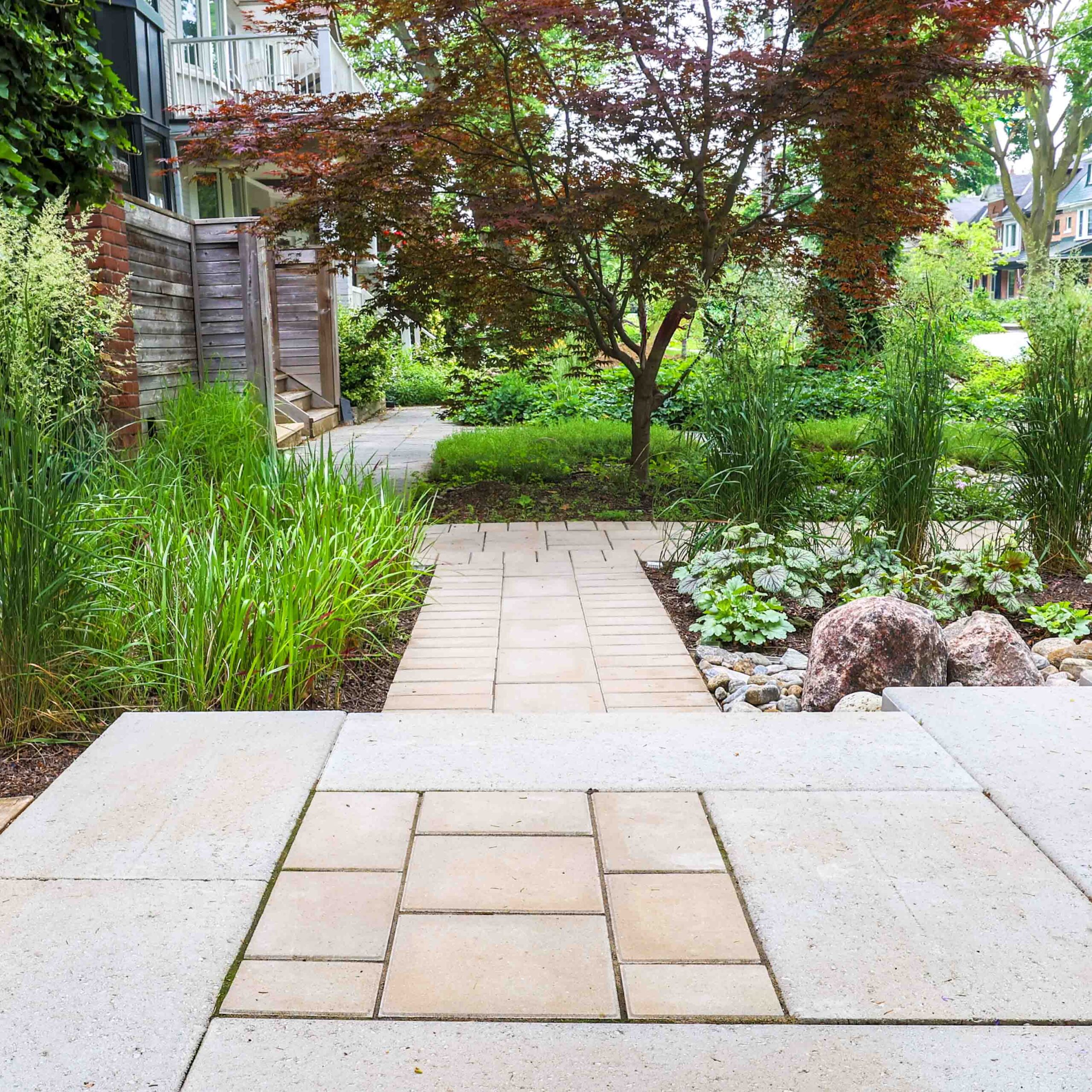 A serene garden pathway bordered by lush greenery, rocks, and a red-leafed tree. The walkway leads towards a building with wooden details.